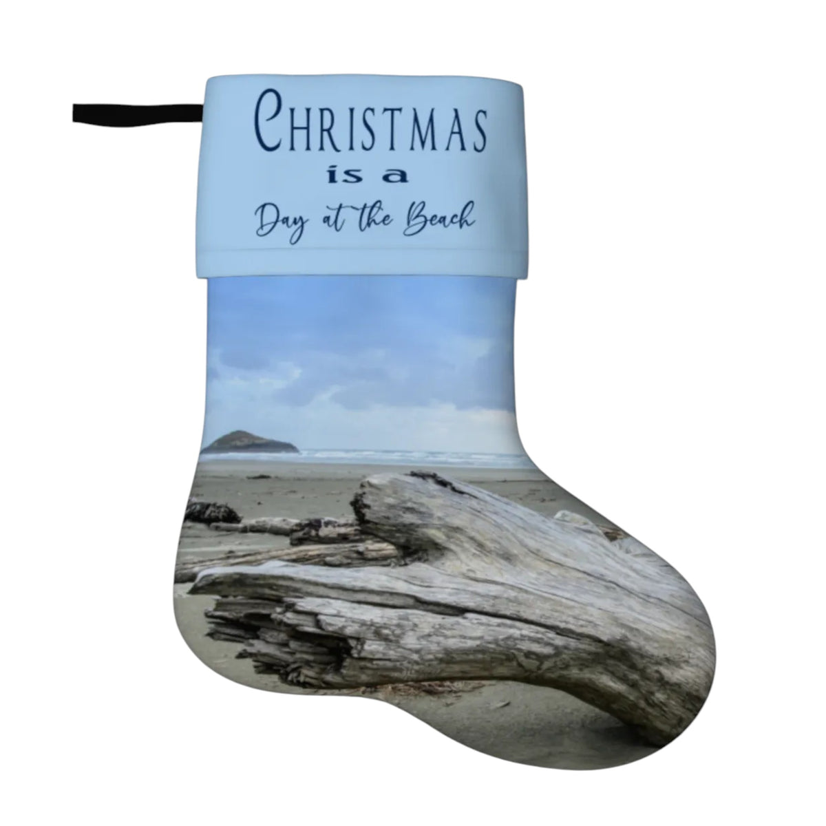 A Christmas stocking with a light blue cuff printed with the text 'Christmas is a Day at the Beach' and a beach scene below featuring sand, ocean, sky, and driftwood, shown on a plain white background.