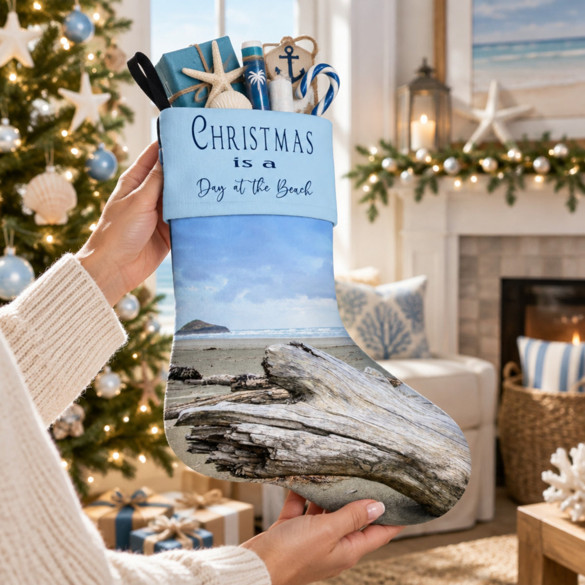 A person holds a Christmas stocking in a cozy living room setting. The stocking features a printed beach scene with driftwood and the text 'CHRISTMAS is a Day at the Beach' on a light blue band.