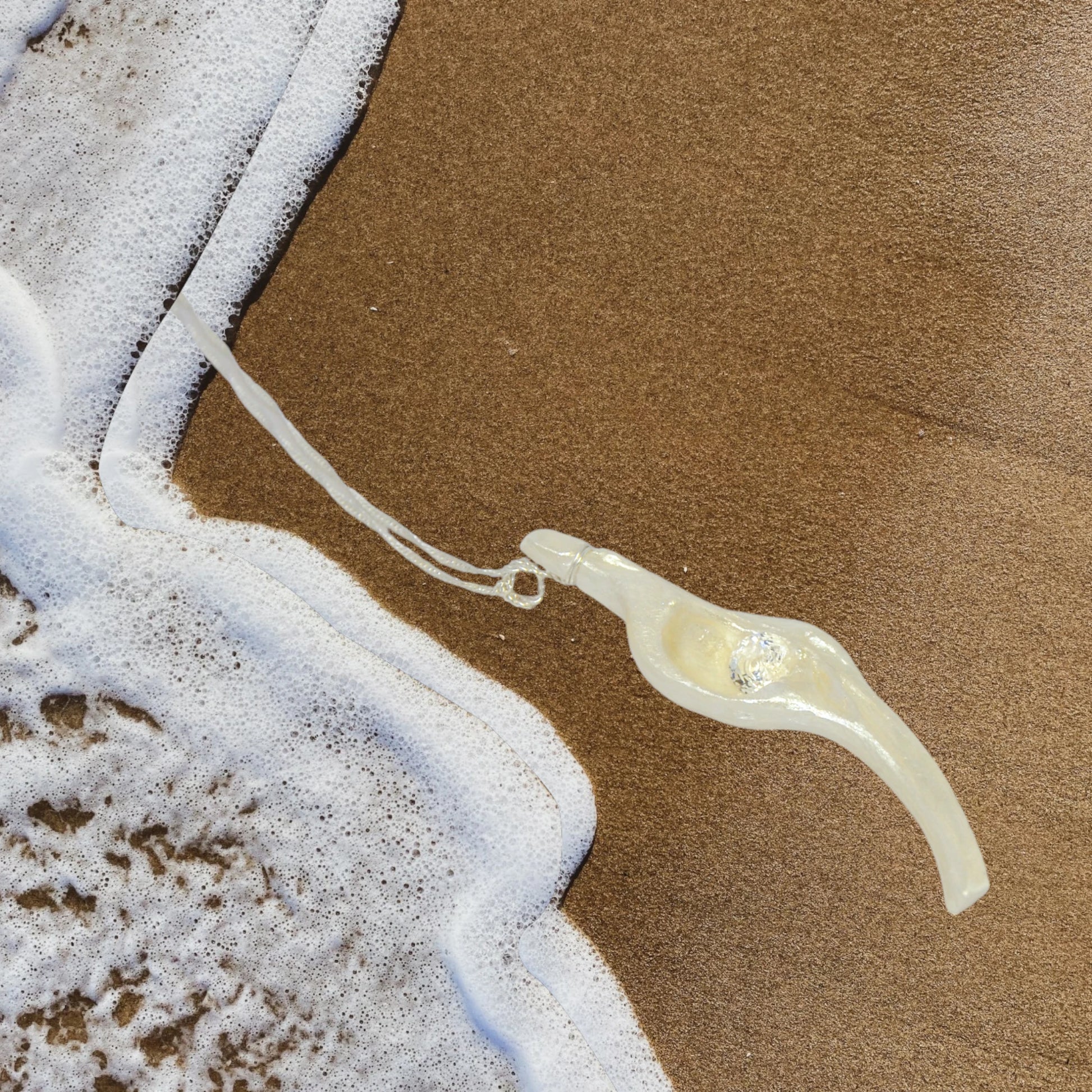 Diamond Star natural seashell pendant with eight mm faceted herkimer diamond. The pendant is laying on the beach sand with ocean waves gently coming up towards the pendant.