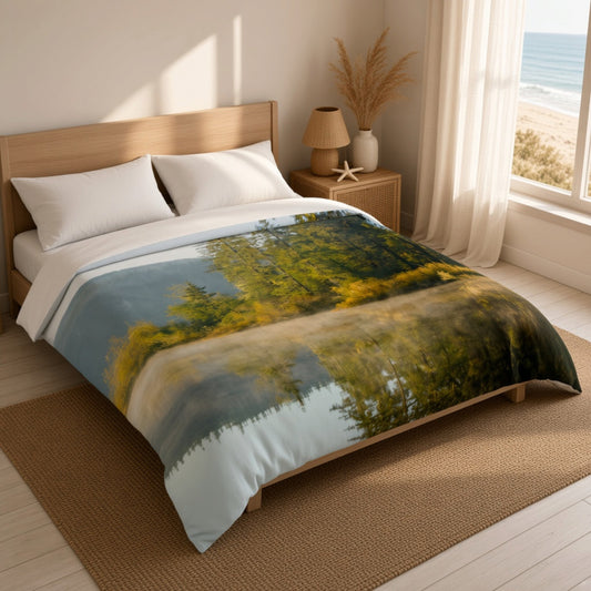 A bed dressed in a scenic landscape duvet cover featuring a misty forest and lake reflection, styled in a bright bedroom with a wooden headboard, bedside lamp, and a window looking out to the beach.