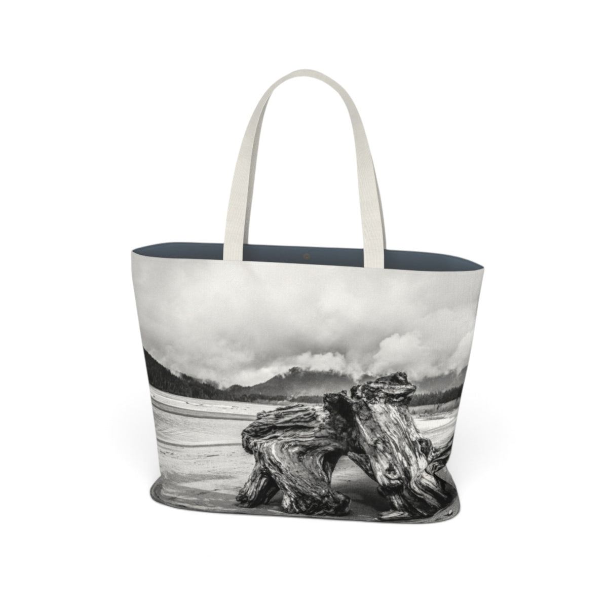 A tote bag featuring a black and white landscape photograph of a driftwood log on a beach, with light beige shoulder straps and a dark navy blue interior lining, shown against a plain white background.
