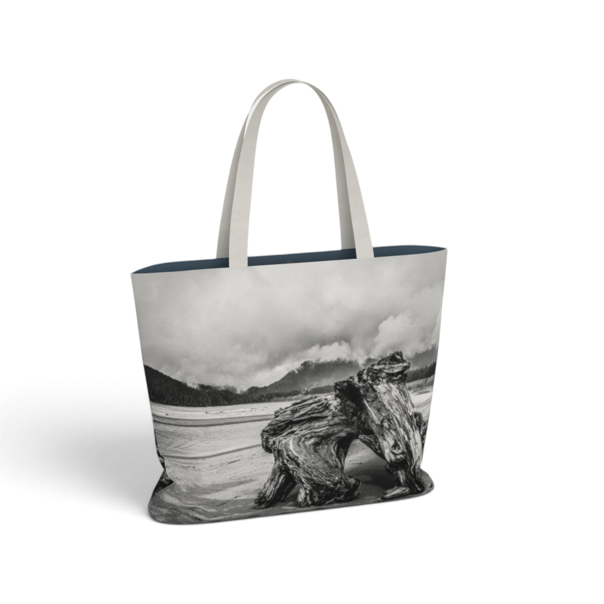 A tote bag featuring a black and white landscape photograph of a beach scene with mountains, clouds, and driftwood, with light gray fabric handles, shown on a white background.