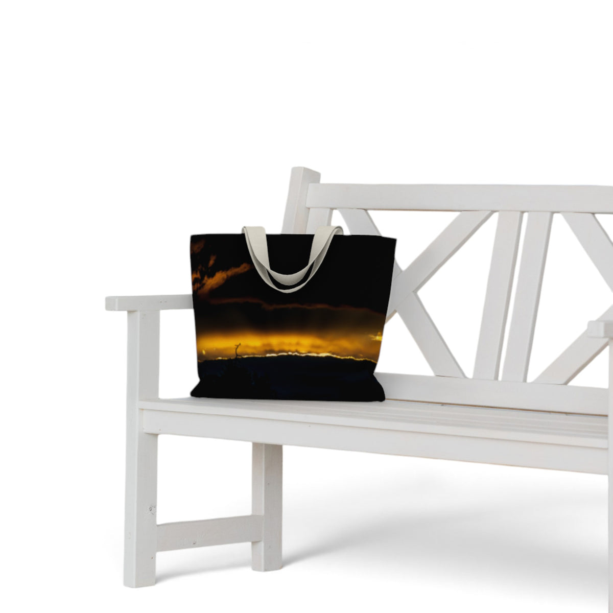 A canvas tote bag with a sunset landscape print sits on a white wooden bench against a white background.