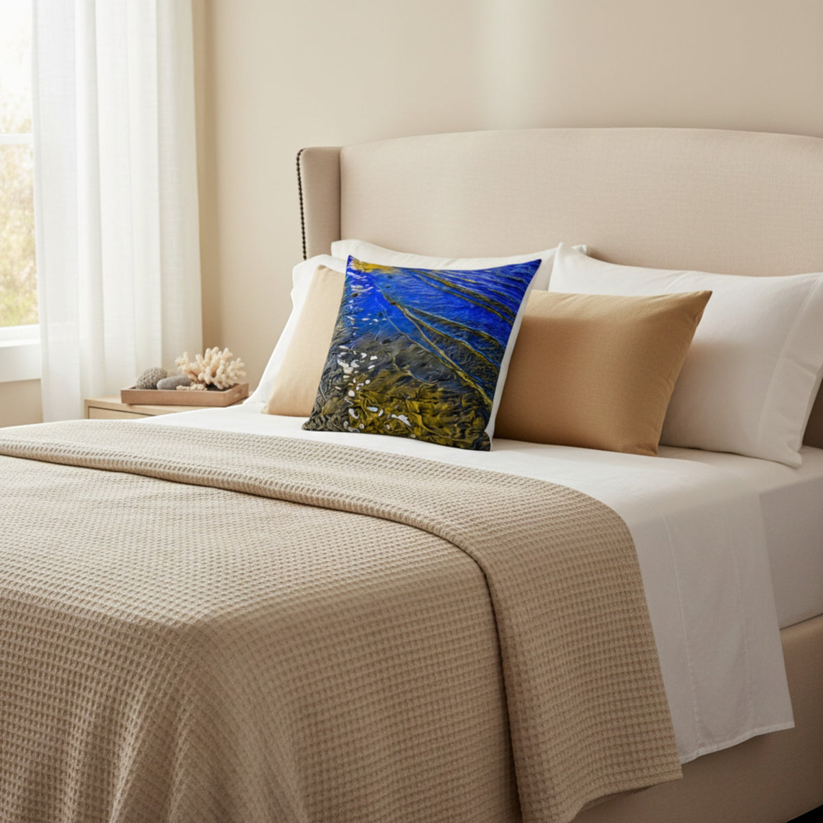 A styled bed in a softly lit bedroom featuring a beige waffle-knit throw blanket, white bedding, and two accent pillows. The most prominent product is a square decorative throw pillow with a vivid blue and green abstract landscape print, set against a neutral beige headboard and wall.