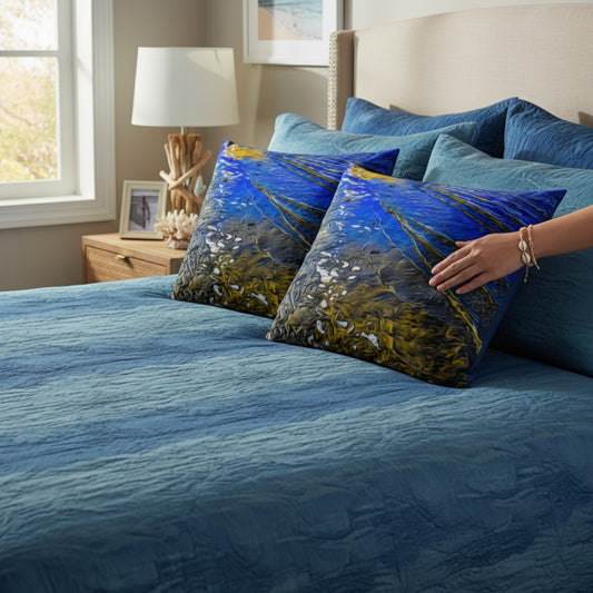 A styled bedroom scene showing a bed with blue bedding and two decorative throw pillows featuring an abstract blue, gold, and white coastal or floral-like print. A person's hand is resting on one pillow.