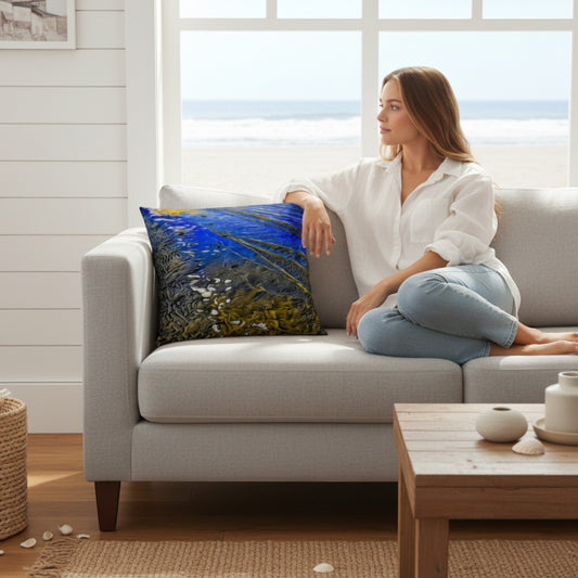 A woman sits on a light gray sofa in a bright living room, looking out a large window toward the ocean. A square throw pillow with a vivid blue and green abstract coastal print rests on the sofa beside her.