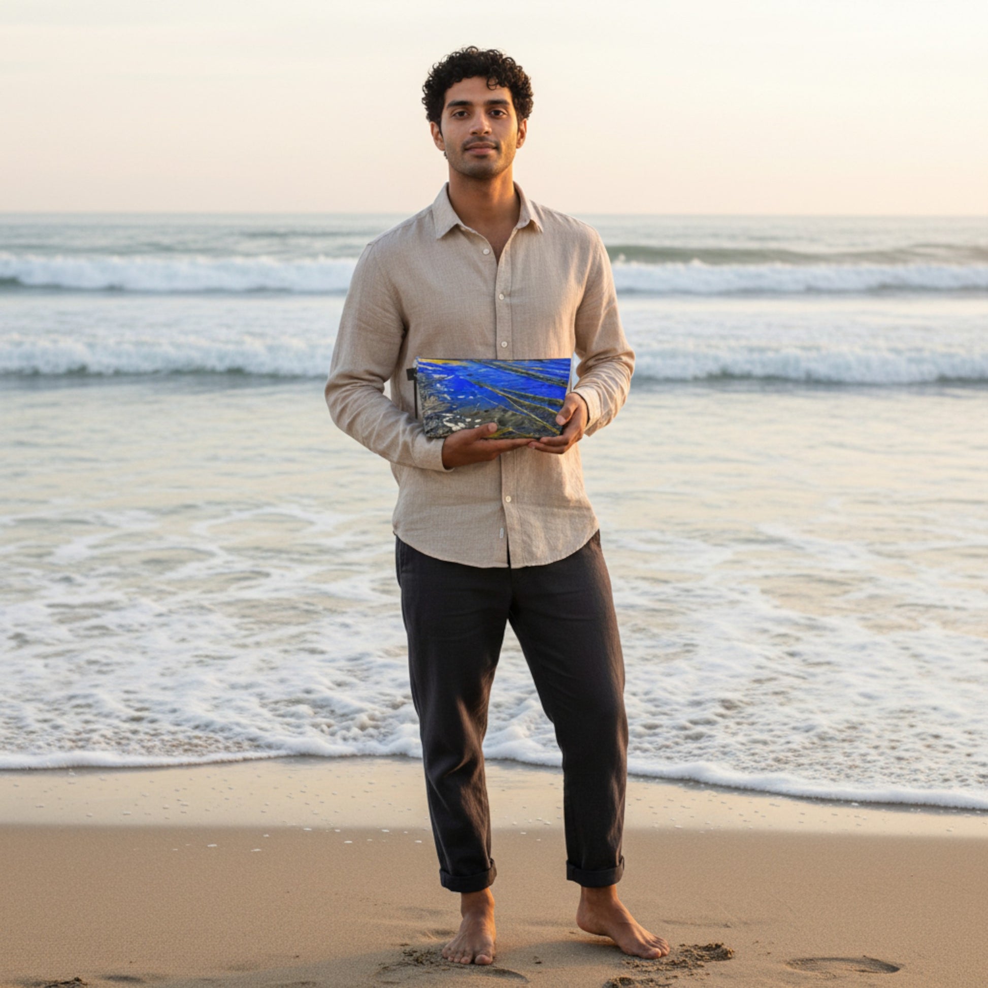 Man holding a blue and yellow travel bag on a beach with ocean waves in the background