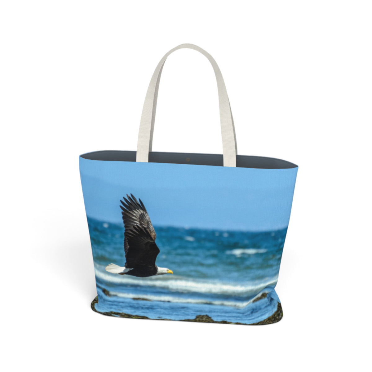 A tote bag featuring a photographic print of a bald eagle flying over blue ocean waves, with light beige fabric handles and a dark navy interior lining, shown against a plain white background.
