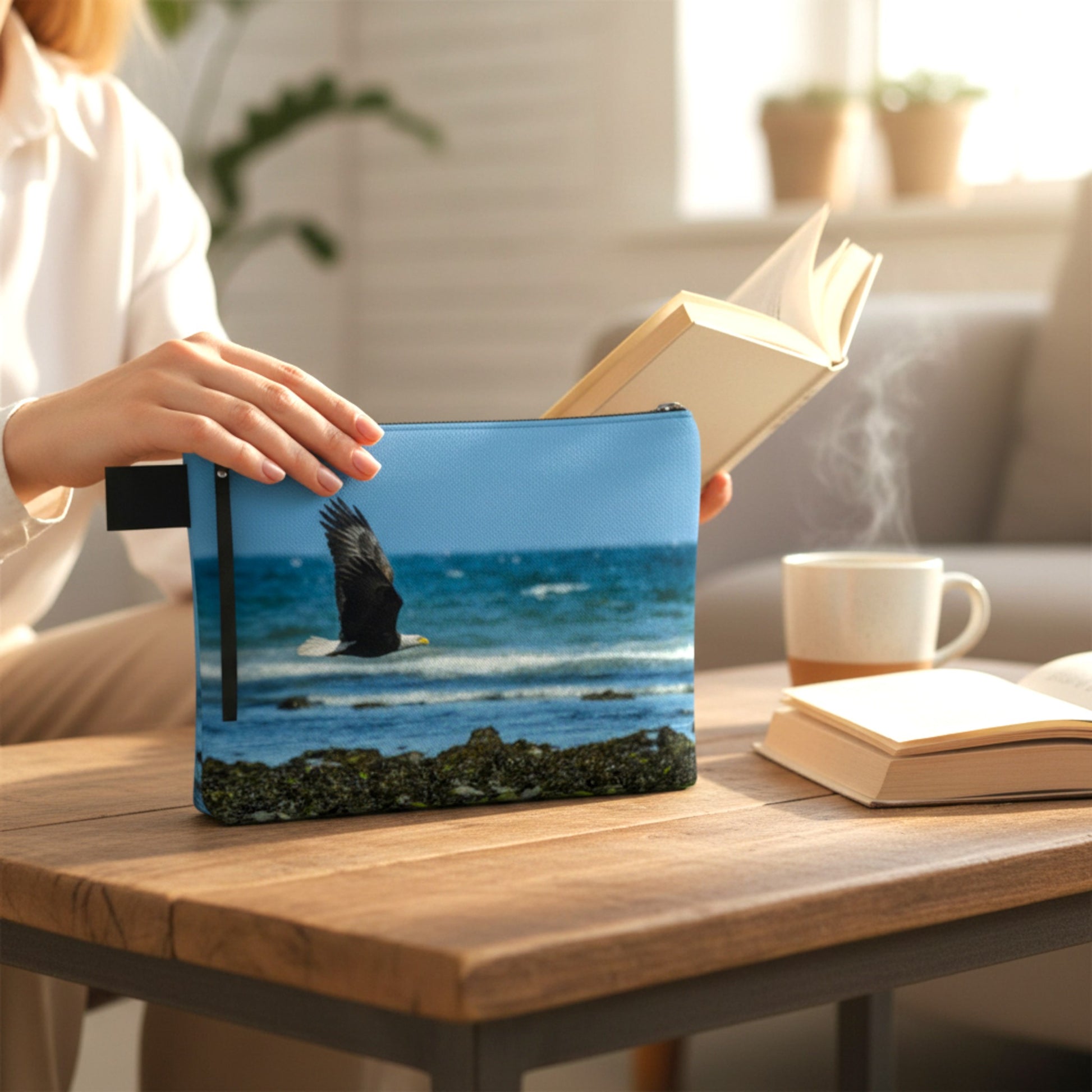 Person holding a pouch with an eagle design on a table with books and a mug.