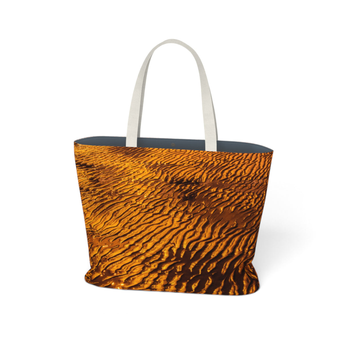 A tote bag featuring a detailed print of golden sand dunes with rippled textures, complete with two white shoulder straps, shown on a plain white background.