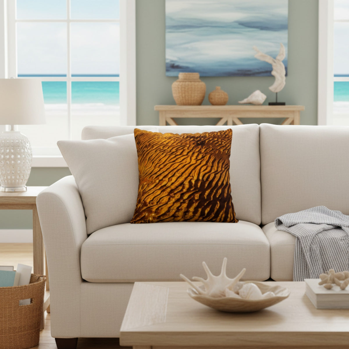 A square throw pillow with a textured golden-brown wavy pattern, styled on a cream-colored sofa in a bright, modern living room with a beach view.
