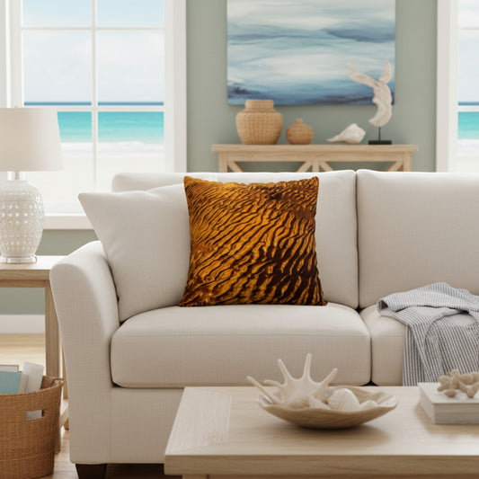 A square throw pillow with a textured golden-brown wavy pattern, styled on a cream-colored sofa in a bright, modern living room with a beach view.
