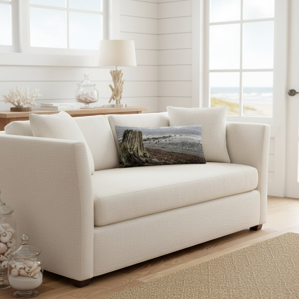 A throw pillow with a beach and ocean scene, featuring a large driftwood log on a sandy beach with a cloudy sky in the background.