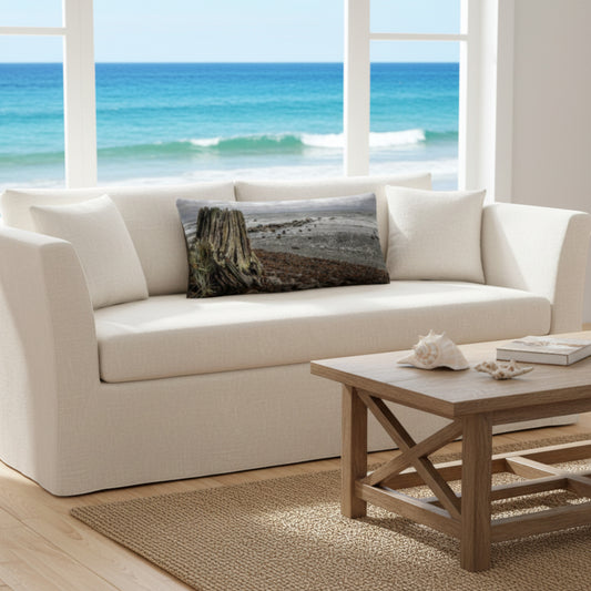 A throw pillow with a beach and ocean scene, featuring a large driftwood log on a sandy beach with a cloudy sky in the background.