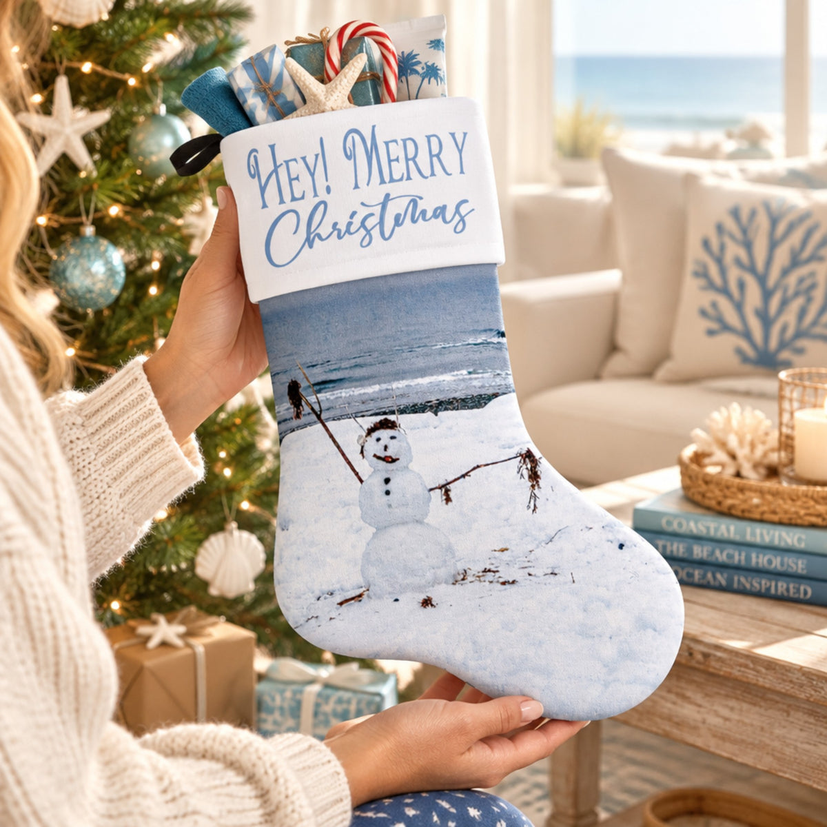 A person holds a Christmas stocking decorated with a winter beach scene and a snowman, featuring a white cuff that reads 'Hey! Merry Christmas.' The background shows a decorated Christmas tree, a cozy living room, and a beach view in the distance.
