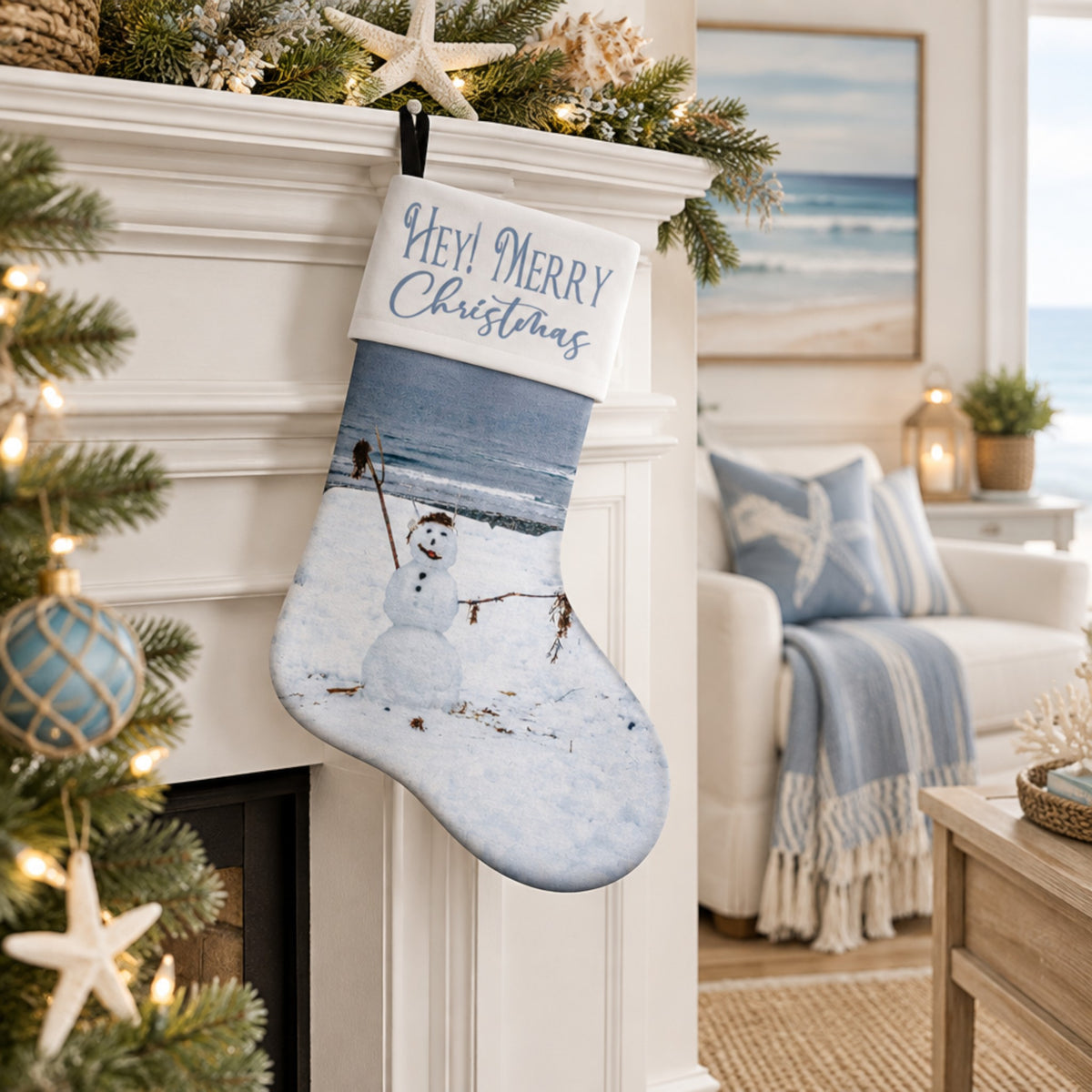 A personalized Christmas stocking with a winter beach scene and the text 'HEY! MERRY Christmas' hangs from a white fireplace mantel decorated with pine branches, a starfish ornament, and a lit Christmas tree. The background shows a cozy living room with a blue-and-white throw blanket and a beach view window.