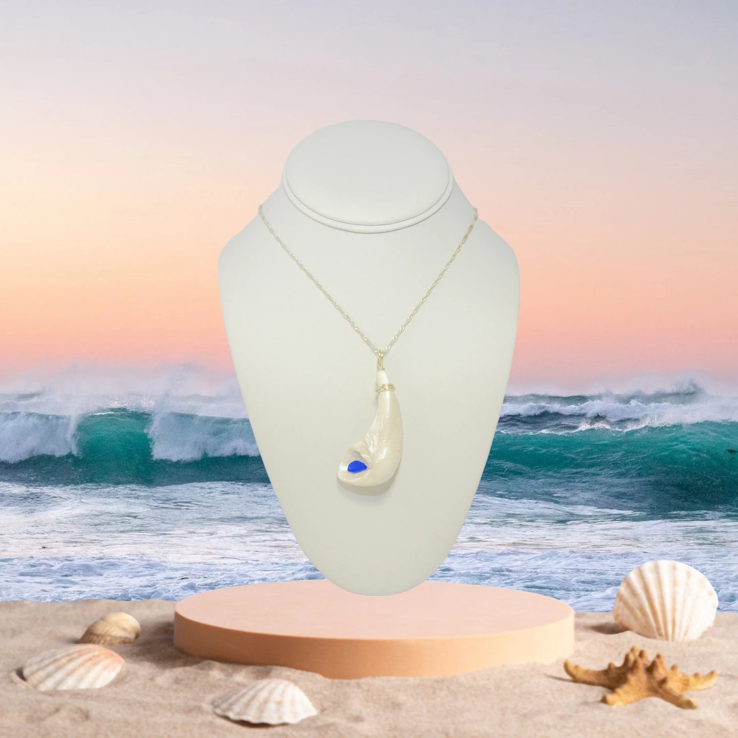The cobalt pendant is shown hanging on a chain on a necklace displayer.  The setting sun over the ocean waves heading towards the sandy beach.  A few seashells and a starfish lay strewn about the sand.