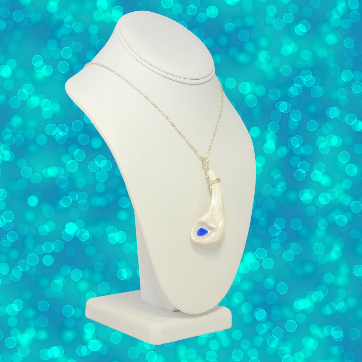The Cobalt pendant is shown hanging from a chain on a necklace displayer.  The pendant is turned to the viewer can see the angle of the pendant when worn. The background is turquoise blue in a bokeh format.