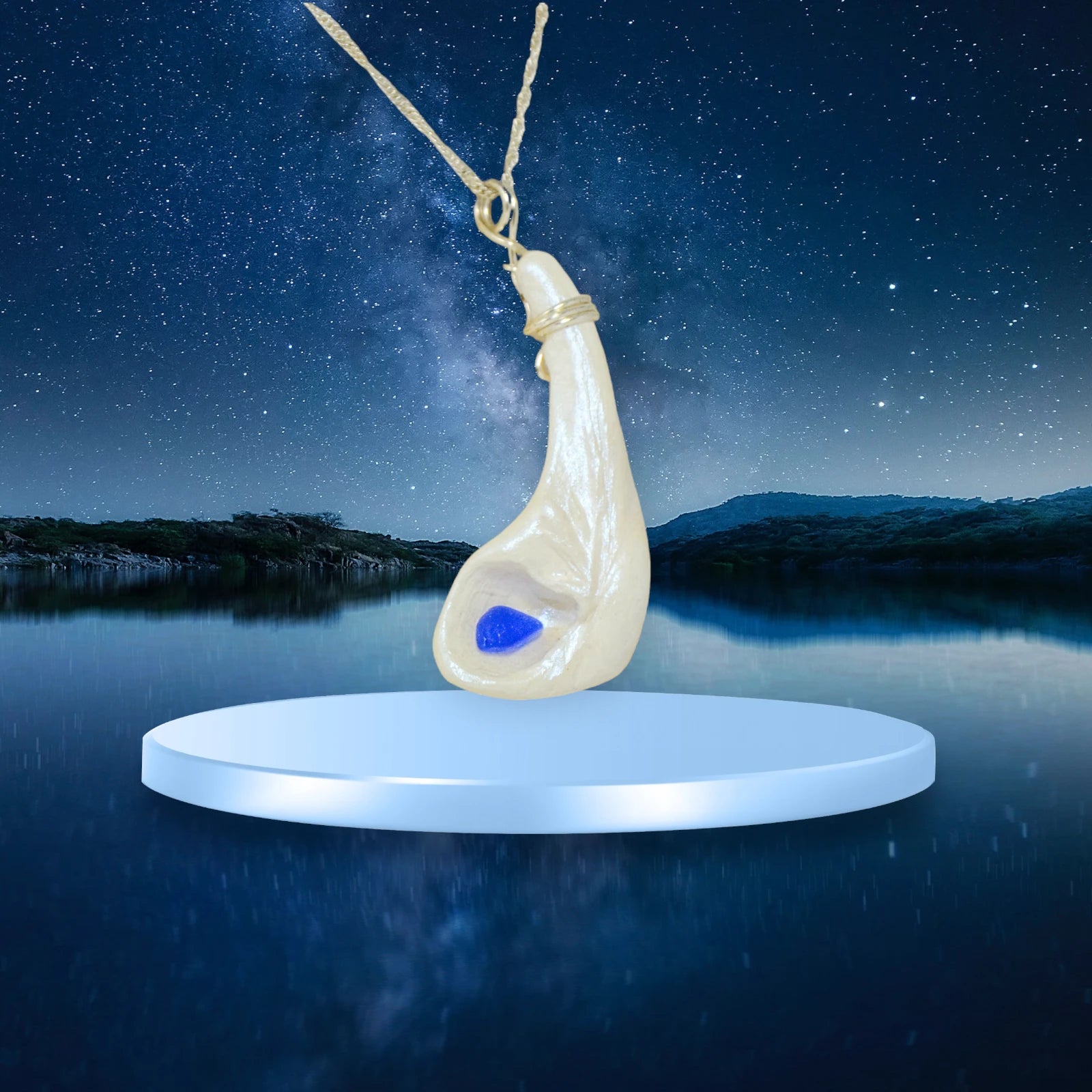 The cobalt pendant with beautiful blue seaglass is shown hanging from a chain with a silvery blue platfrom over a lake with a night sky.  The milky way sits behind the pendant.