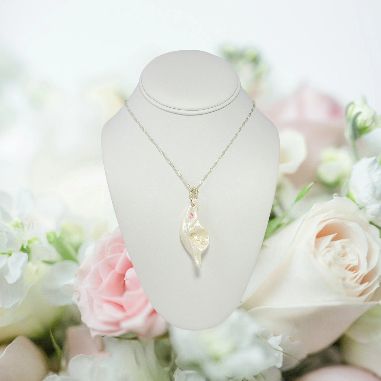 A trio of white baby freshwater pearls and a teardrop rose cut pink tourmaline gemstone compliments the seashell pendant beautifully. The pendant is shown hanging on a necklace over a white displayer with pink and white roses in the background.