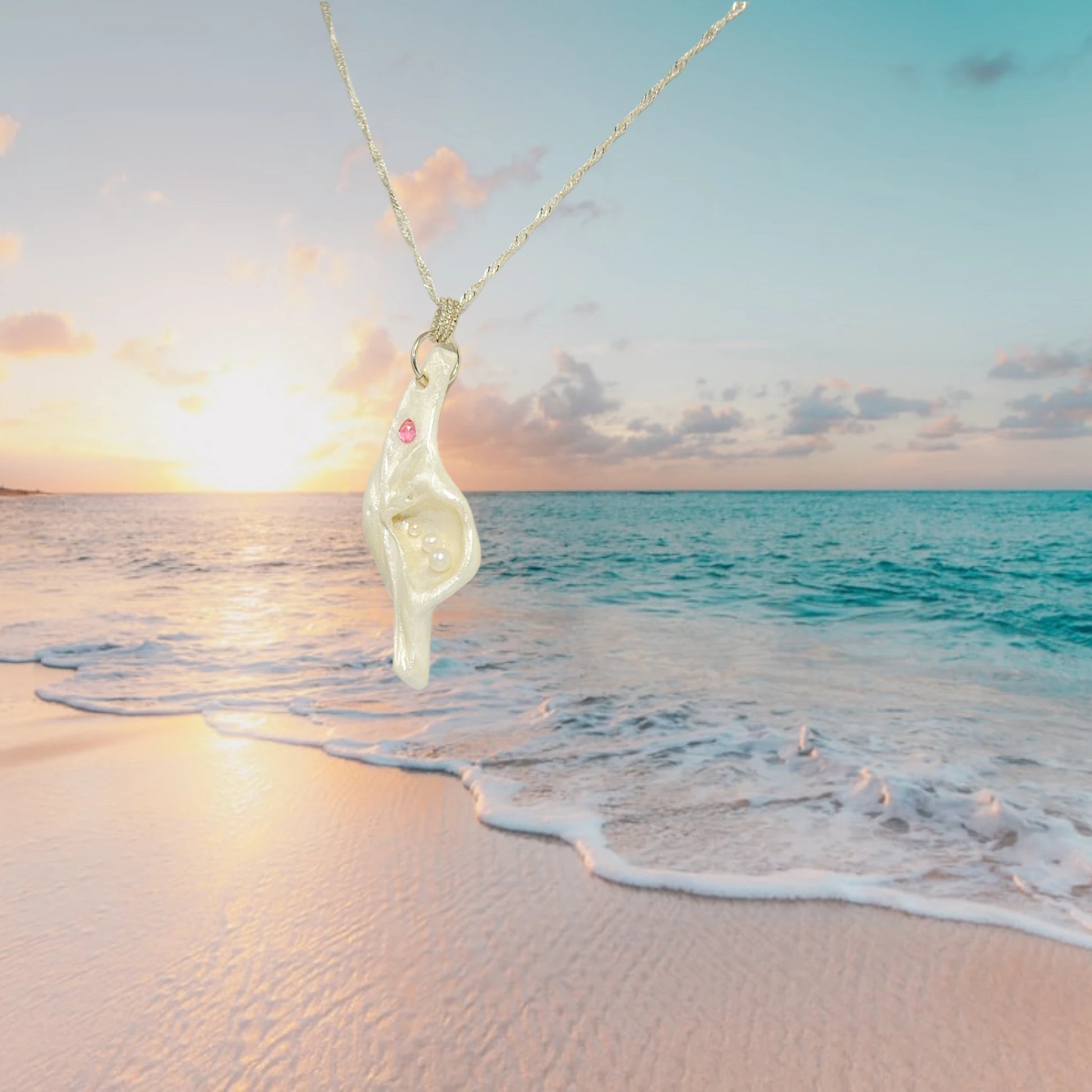 A trio of white baby freshwater pearls and a teardrop rose cut pink tourmaline gemstone compliments the seashell pendant beautifully. The pendant is hanging on a chain with a beautiful sunrise over the ocean in the background.