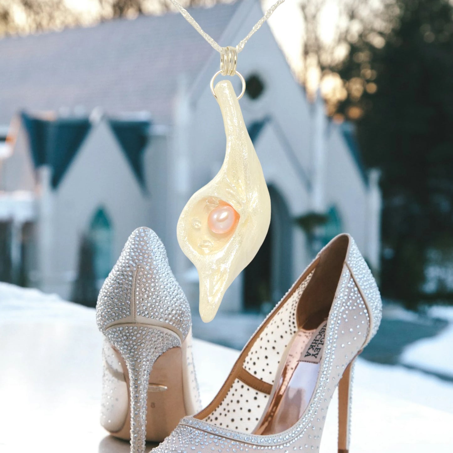 The pendant champagne on ice is shown hanging from a chain.  A church is blurred in the backgroun.  A fancy pair of rhinestone studded high heels are beneath the pendant.  Wedding vibes. van isle goddess dot com