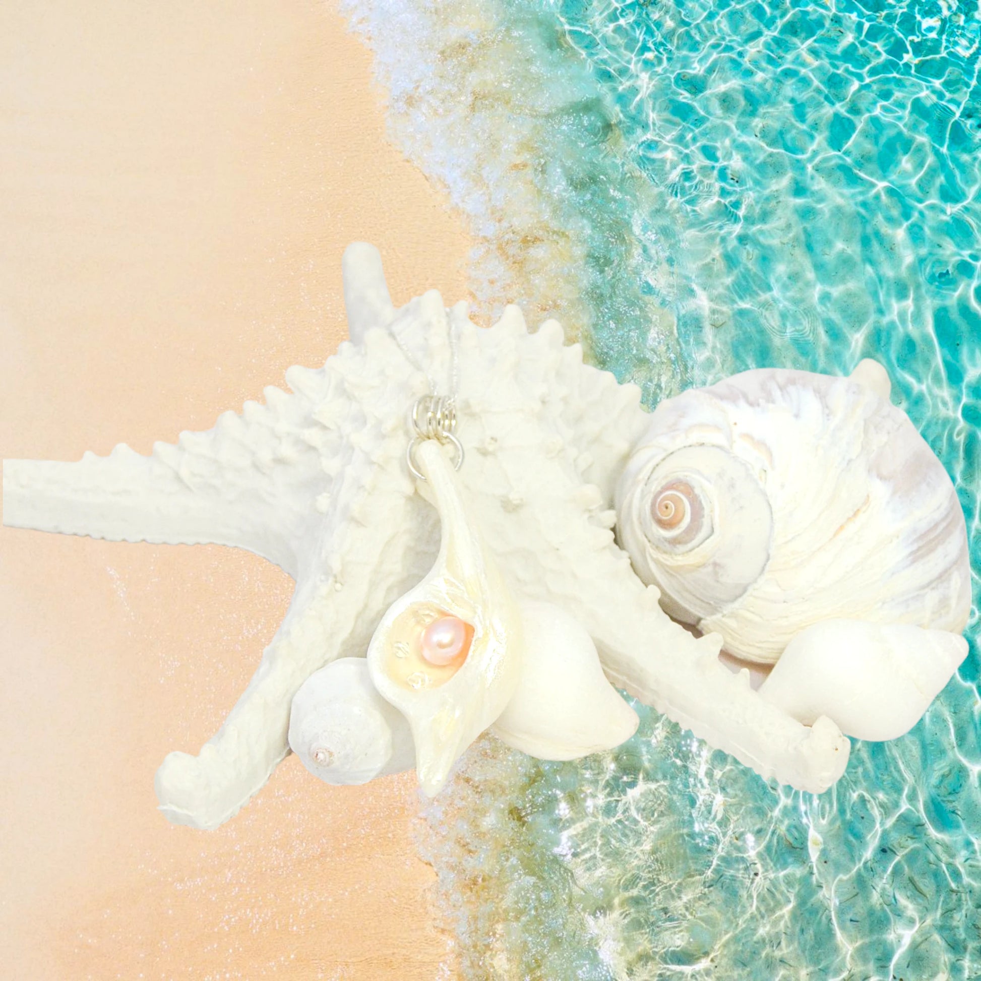 The natural seashell pendant Champagne on ice is shown resting on several smaller seashells and a starfish and a moon snail shell.  The background is the sand and ocean.  Summer vibes.