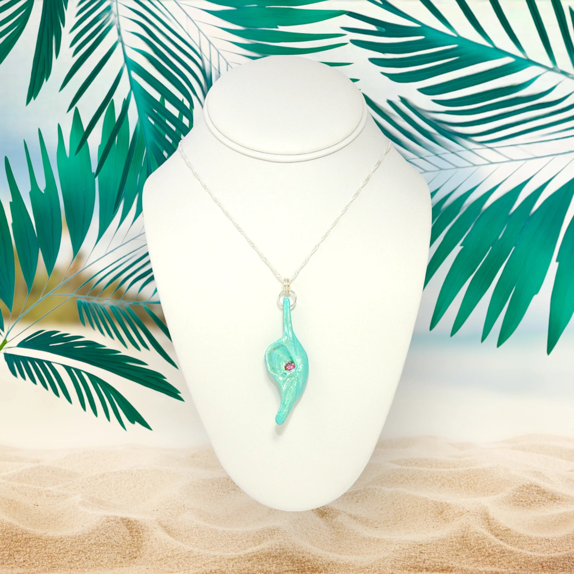 Rio is a natural seashell pendant with a faceted Pink Tourmaline gemstone. The pendant is shown hanging on a white necklace displayer on the sandy beach surrounded by greenery.