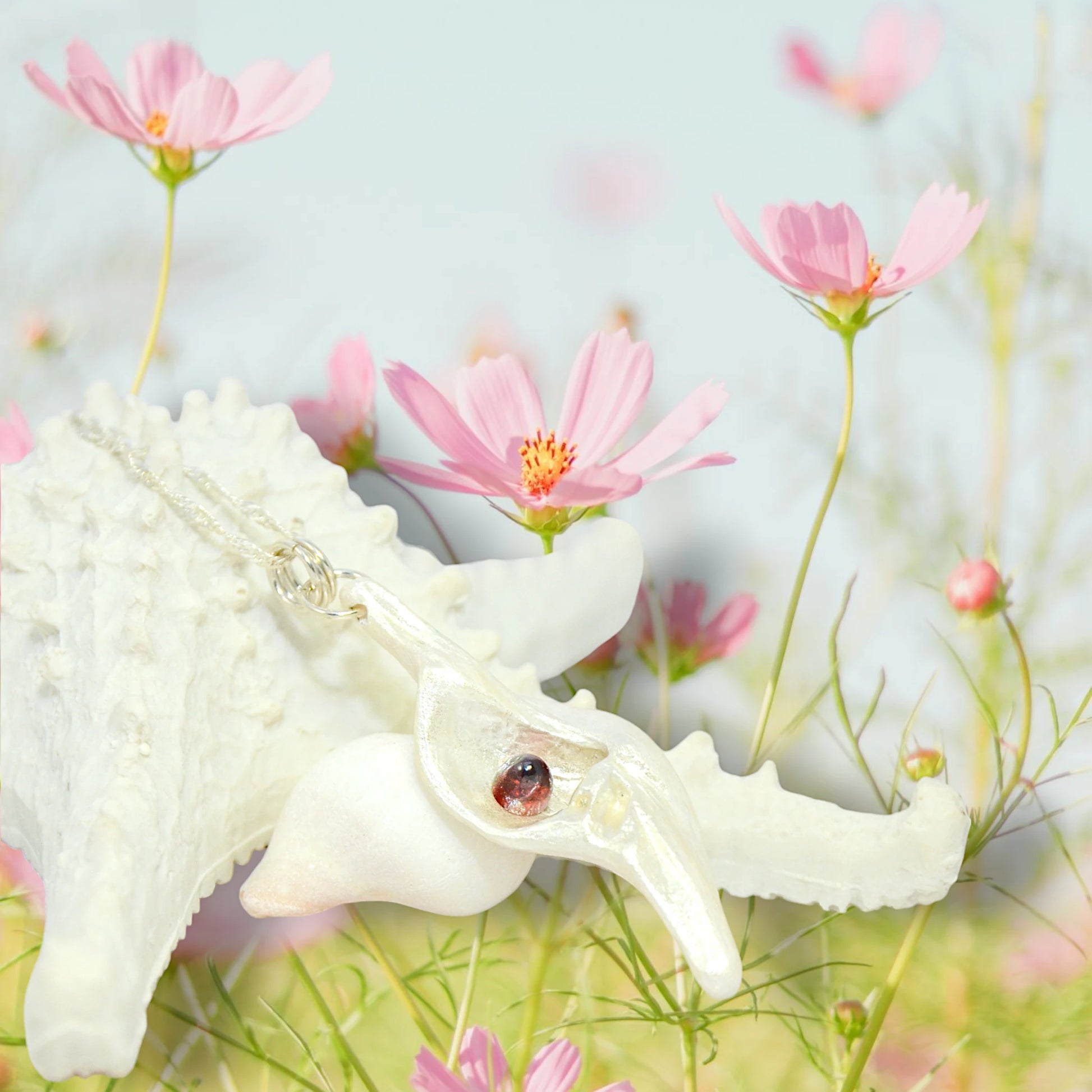 Pomegranate natural seashell pendant is adorned with a stunning Garnet gemstone. The pendant is resting on starfish in a field of pink cosmo flowers.