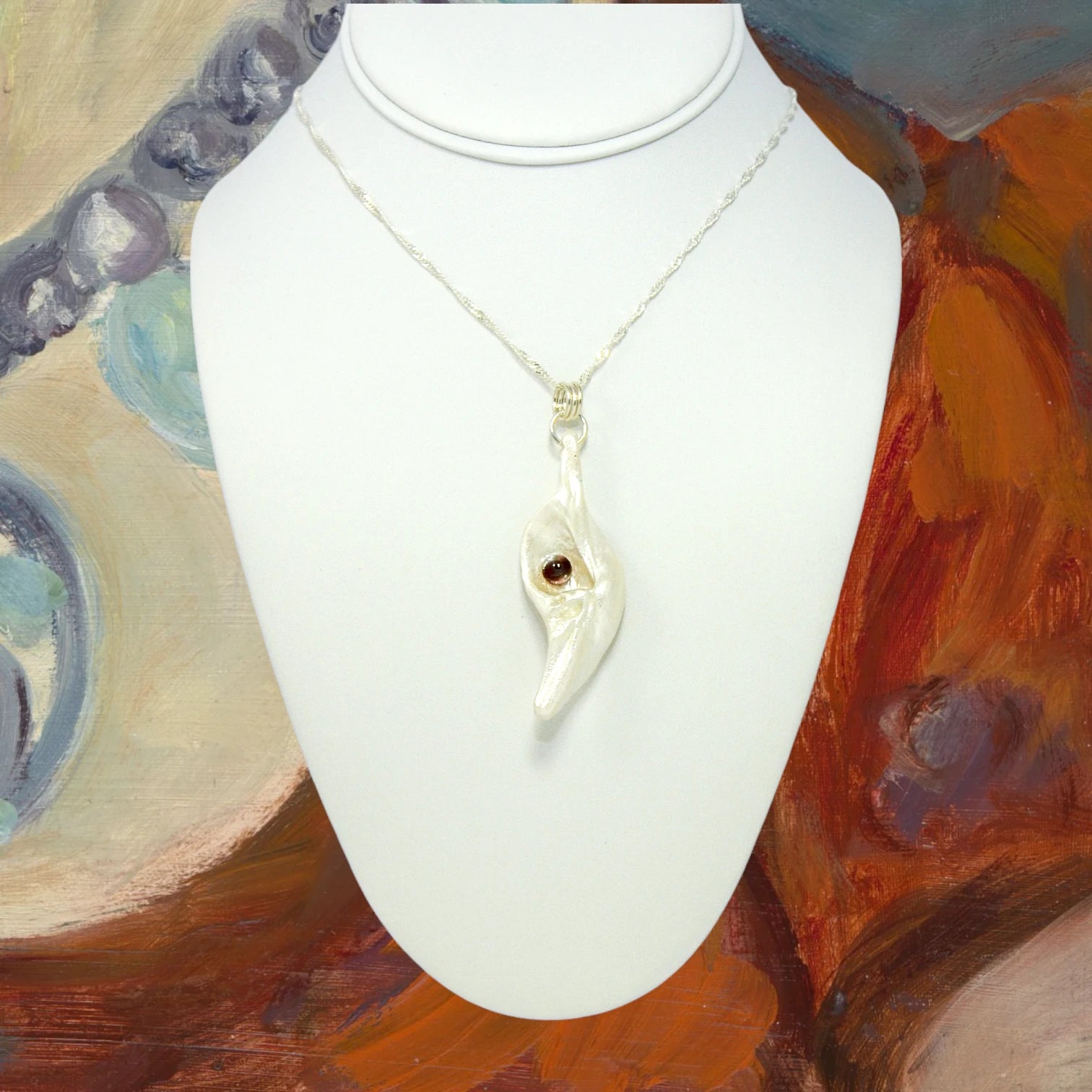 Pomegranate natural seashell pendant is adorned with a stunning Garnet gemstone. The pendant is shown on a white necklace displayer .  The background is of a painting.