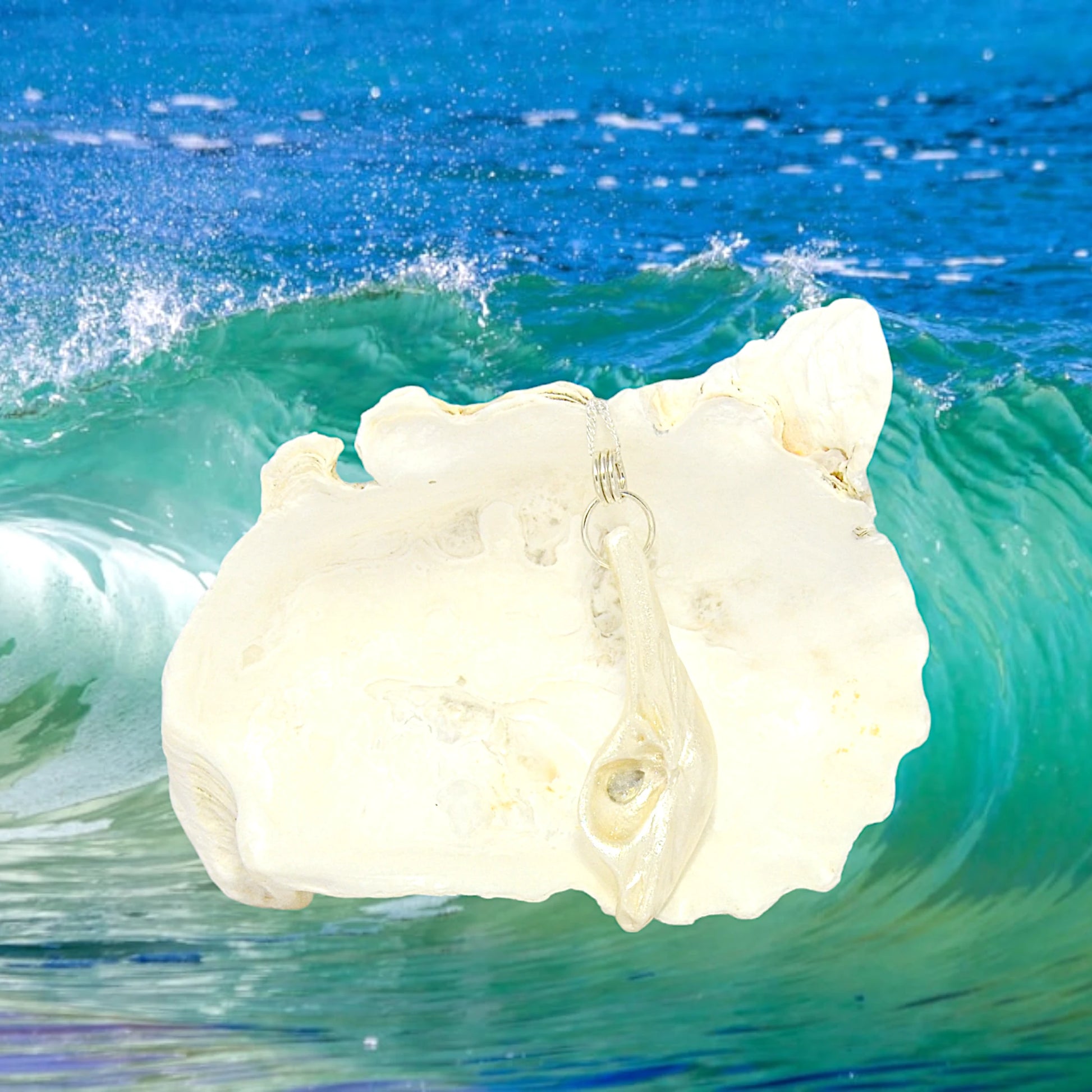   Is shown hanging from a chain with a soft blue floral background. The pendant is shown hanging from a larger seashell with ocean waves in the background.