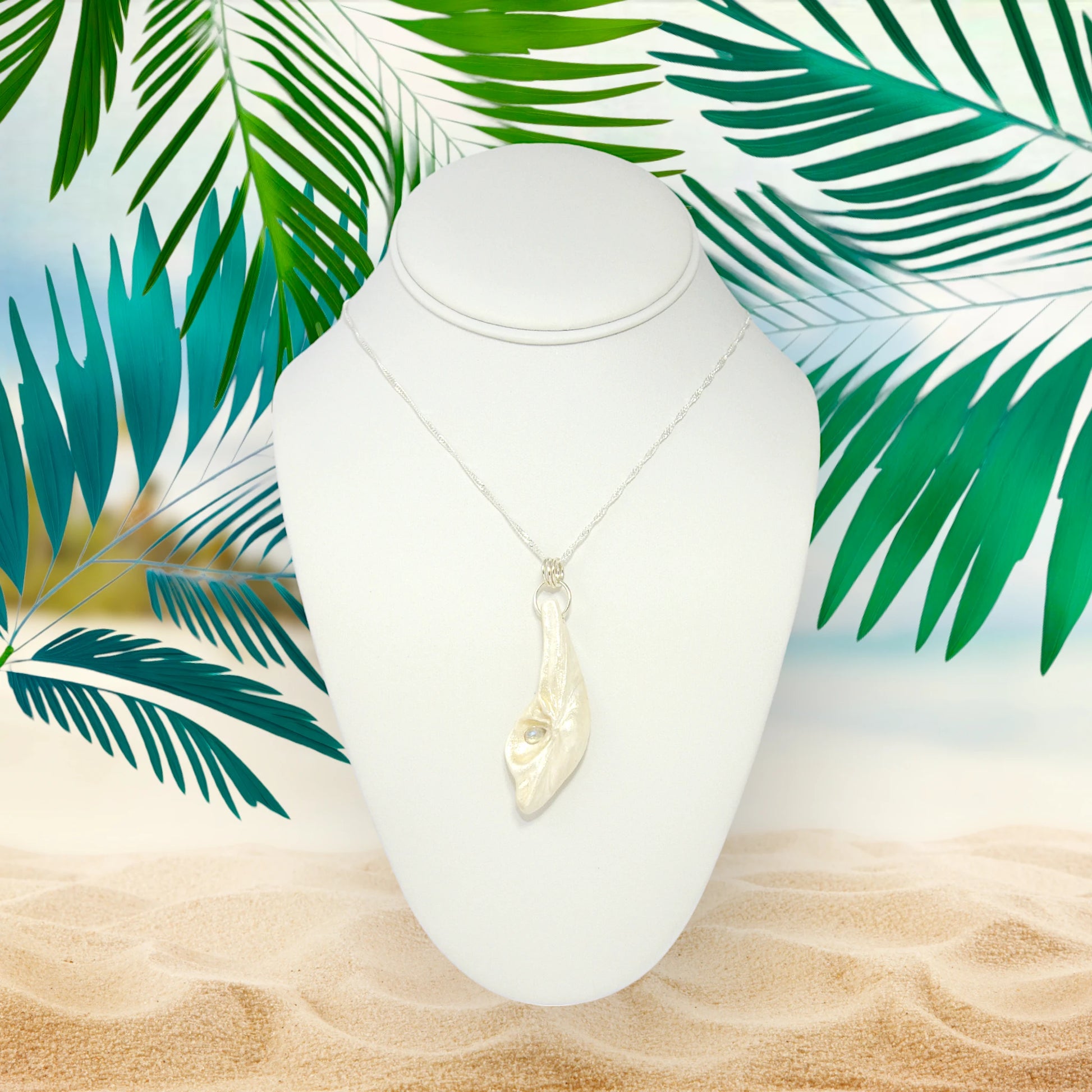 Cool Summer pendant made of natural seashell with a tear drop shape rose cut labradorite gemstone. The pendant is shown on a white necklace displayer.  The background is a sandy beach with greenery behind the display.