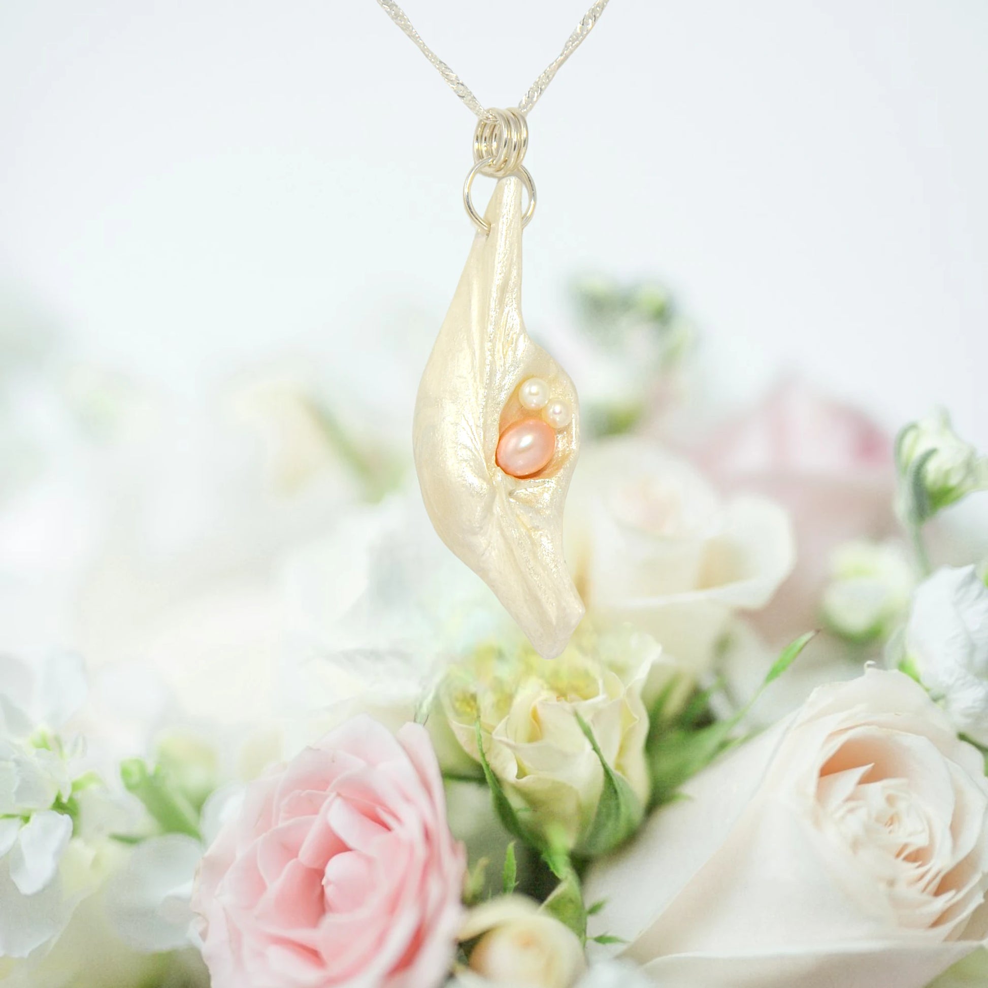 This natural seashell pendant has a real 7-8mm pink freshwater pearl and two baby pearls. The pendant is shown hanging from a chain over top a bouquet of pink and white roses.