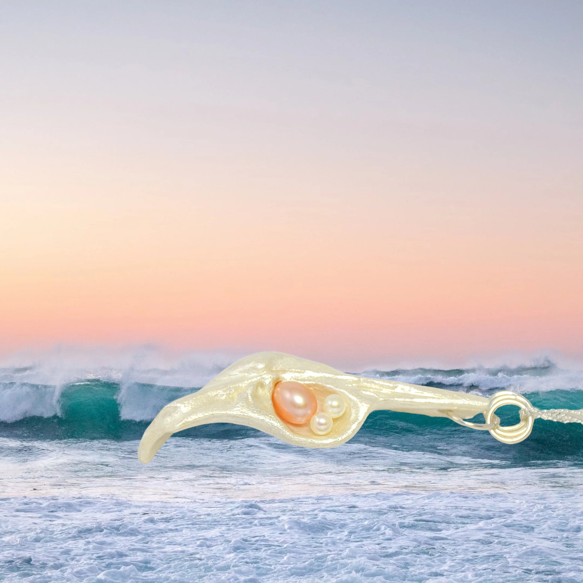 This natural seashell pendant has a real 7-8mm pink freshwater pearl and two baby pearls. The pendant is shown on its side with the ocean waves behind it.