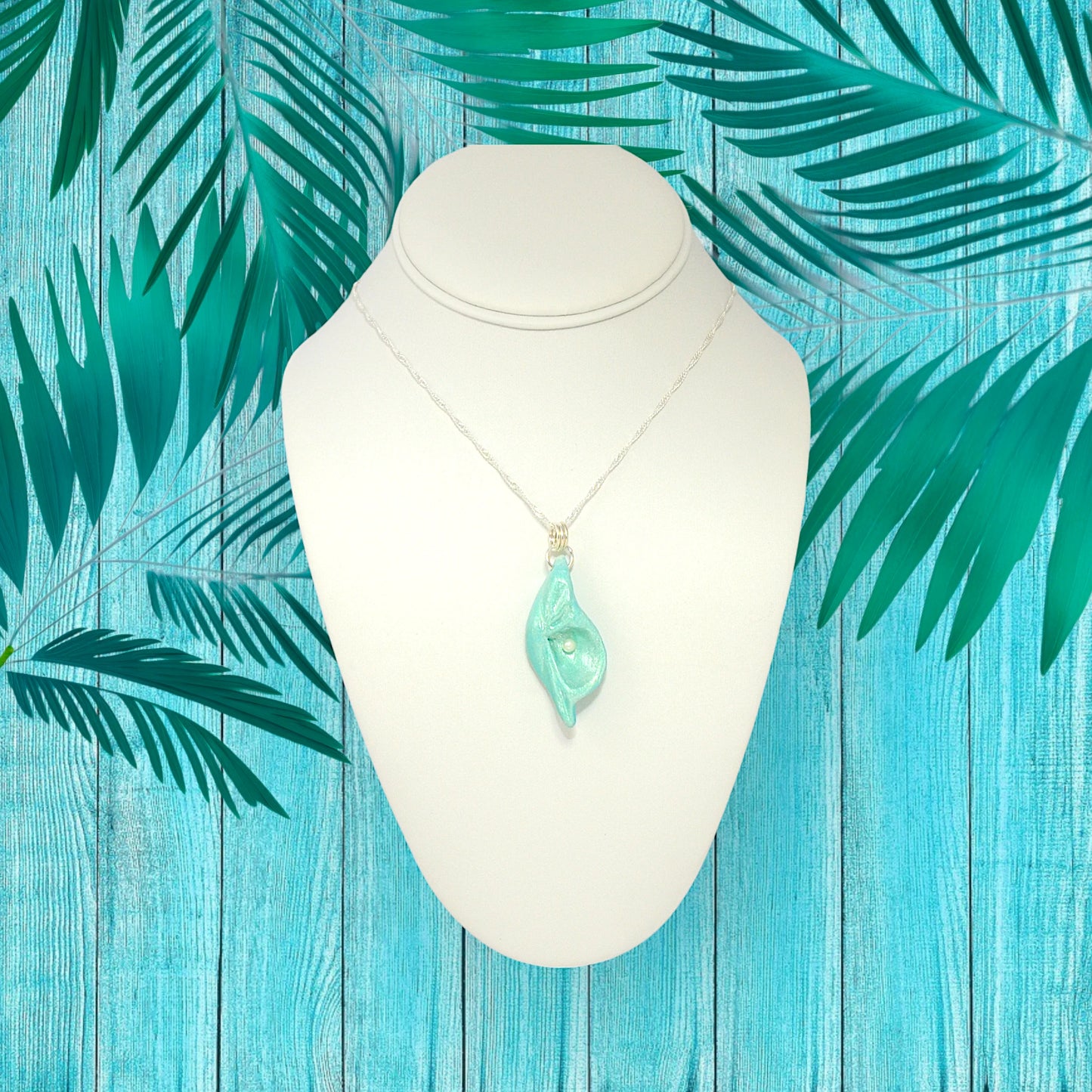 Ocean Pearl natural seashell pendant has a  real freshwater baby pearl. The pendant is shown on a white necklace displayer with a blue fence and greenery.