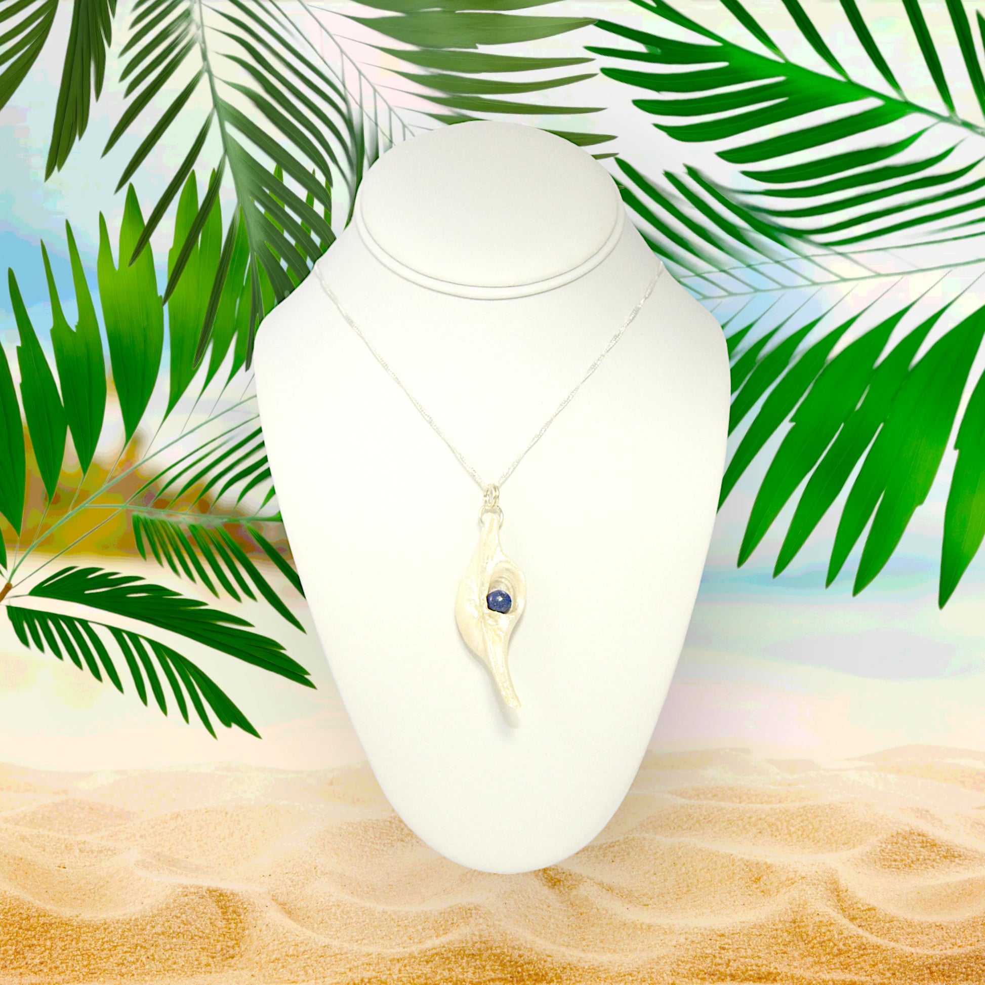 Vision is a natural seashell pendant with a Lapis Lazuli gemstone.  The pendant is shown on a white necklace displayer on a sandy beach surrounded by greenery.