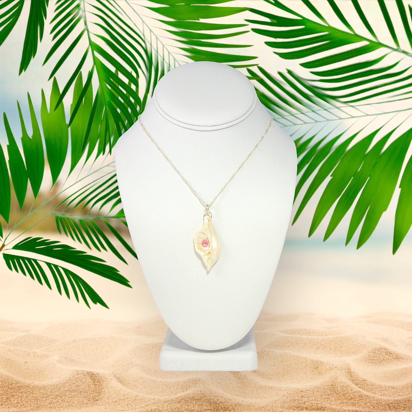 This natural seashell pendant has Pink Tourmaline gemstone and three Herkimer Diamonds that compliments the pendant. The pendant is shown on a white necklace displayer on the sandy beach with greenery.