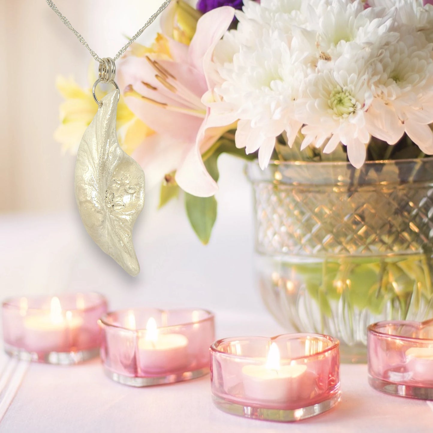 Beautiful pendant called Clarity.  Clarity is a natural seashell from the beach of Vancouver island with seven herkimer diamonds. The pendant hangs from a chain on the left side of the image.  The background shows a bouquet of white mums in a crystal bowl on the right side of the image.  Several pink heart shaped tealight holders with lit tealight candles are on a table below the pendant.