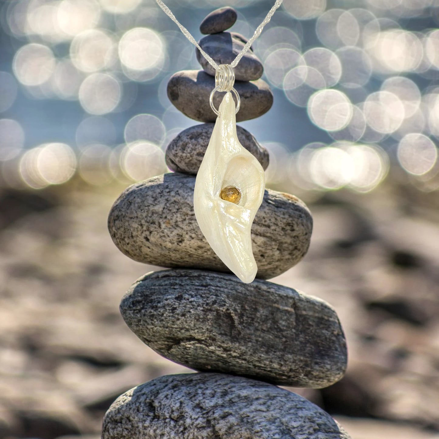 Seer is a natural seashell pendant with a beautiful 5mm rose cut Smokey Quartz compliments the pendant. The pendant is hanging over several rocks that have been stacked one on top of the other.