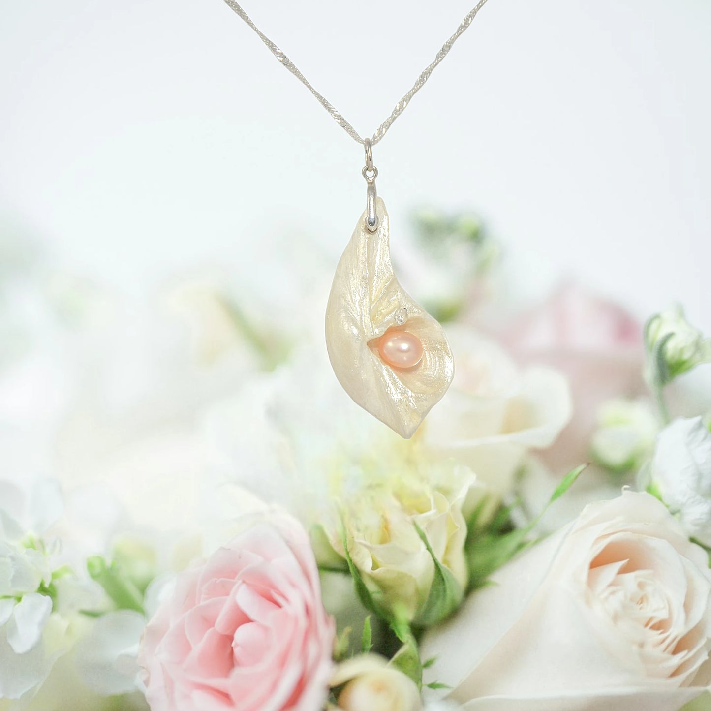 Athena is the name of the pendant being showcased.  It is a natural seashell from the beaches of Vancouver Island. The pendant has a real pink freshwater pearl and a faceted herkimer diamond. The pendant is hanging over a bouquet of pink and white roses.