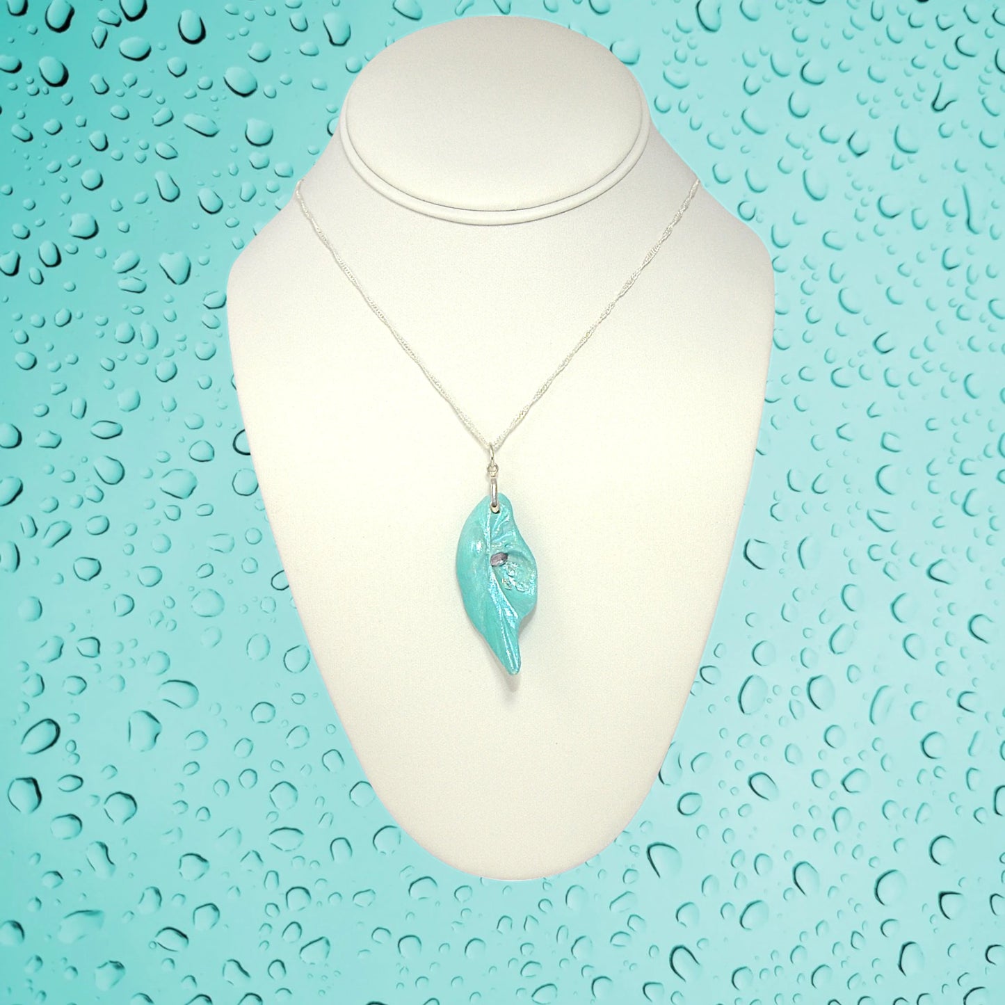 Violet Light is a natural seashell pendant with a rose cut Amethyst Gemstone and three Herkimer diamonds. The pendant is shown on a white necklace displayer with water drops on a turquoise background.