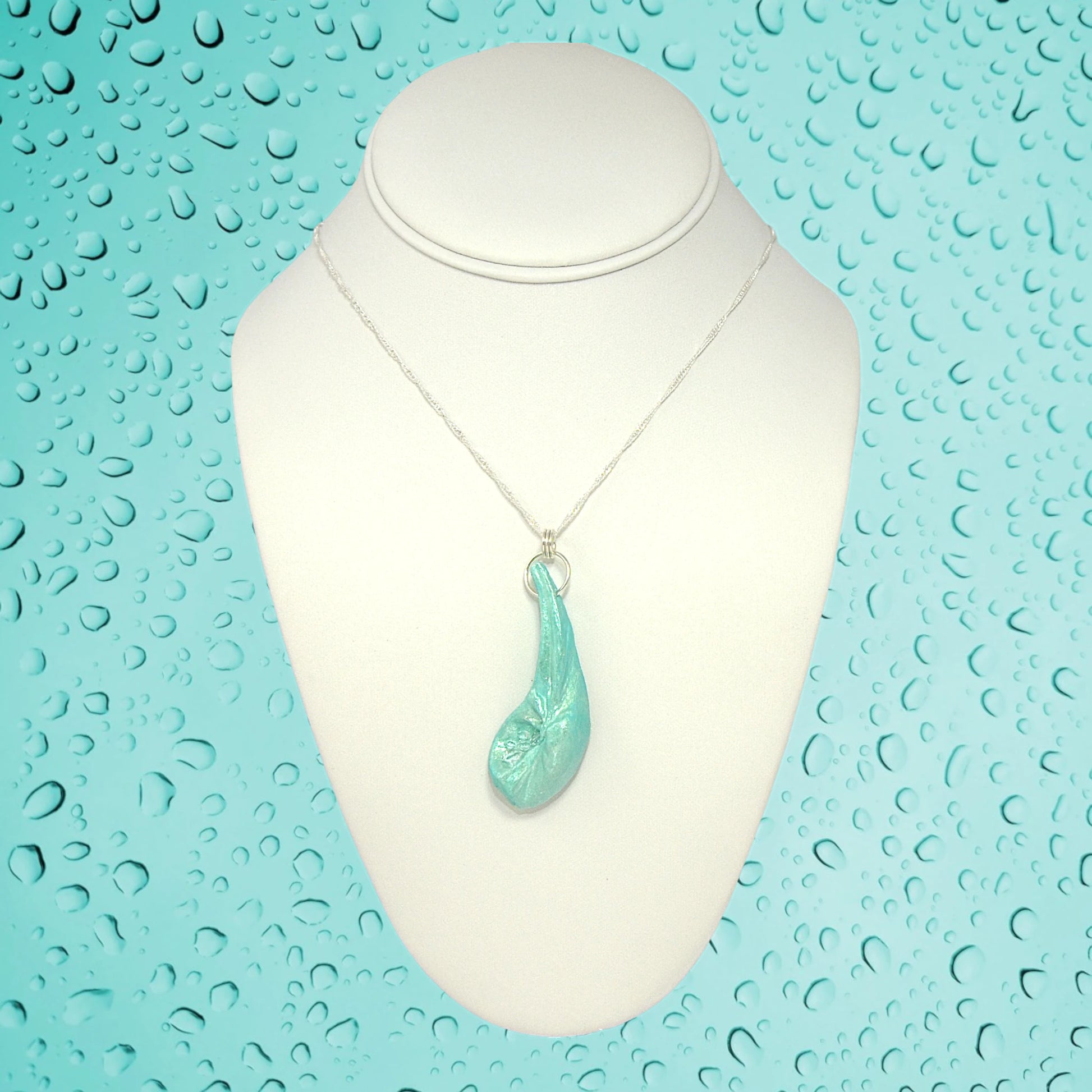 The Cascadia Pendant created with a natural seashell from the beaches of Vancouver Island.  The seashell is turquoise and has high quality herkimer diamonds. The pendant is hanging from a chain on a white necklace displayer.  The background is a light turquoise colour to compliment the colour of the pendant. There are rain drops on the turquoise background.