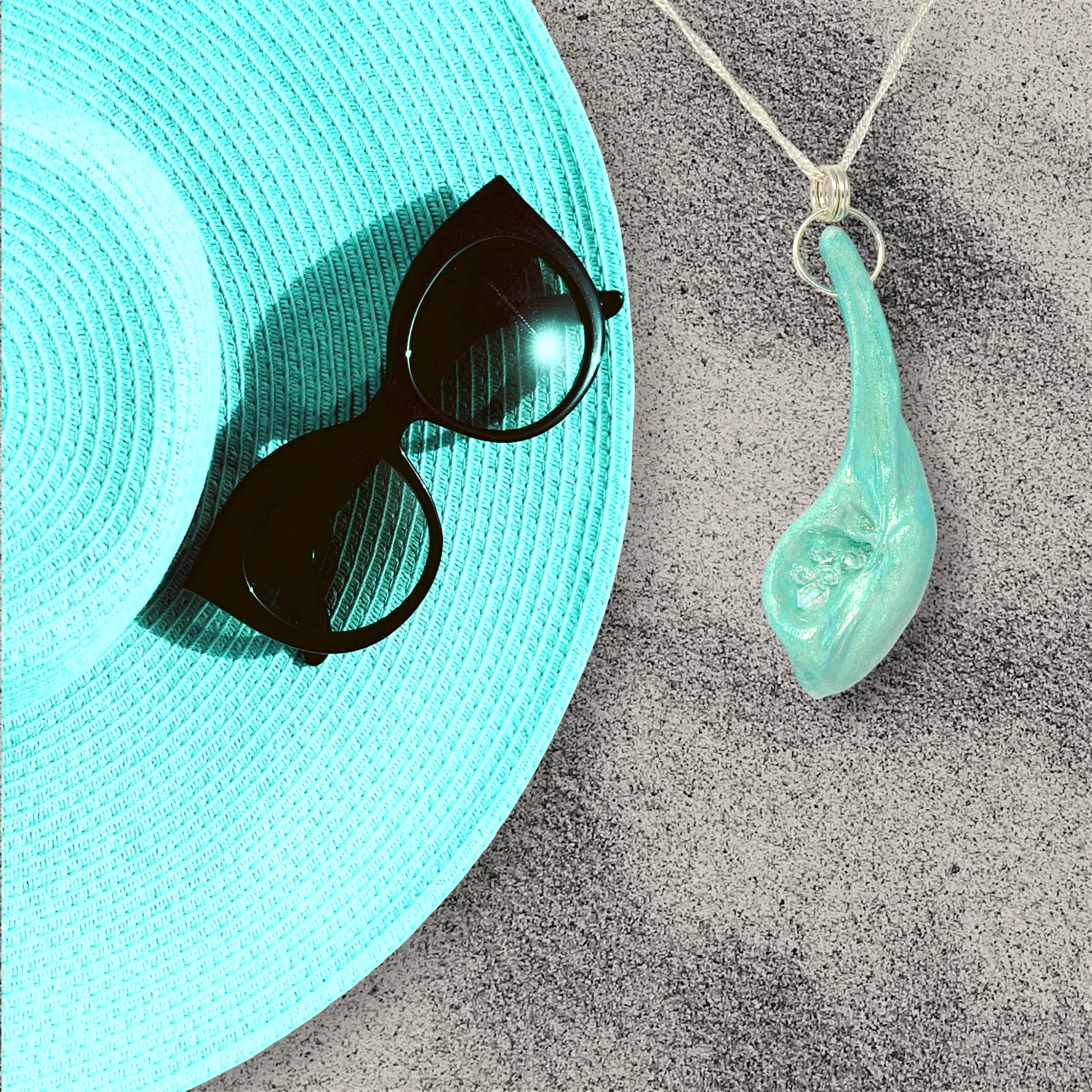 The Cascadia Pendant created with a natural seashell from the beaches of Vancouver Island.  The seashell is turquoise and has high quality herkimer diamonds.  The pendant is shown laying on the sand beside a turquoise summer wide brimmed lades had and black sunglasses.