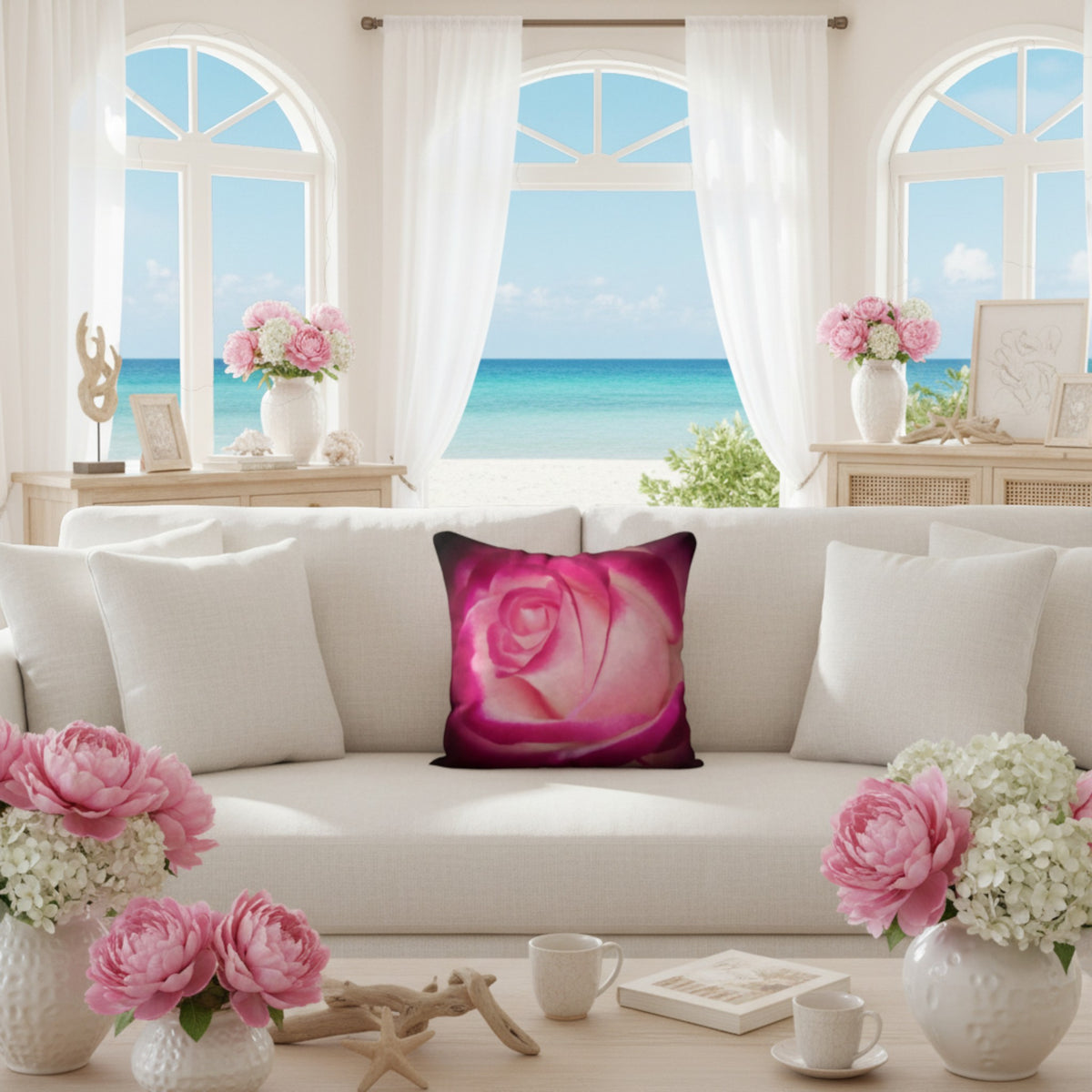 A square throw pillow featuring a close-up print of a pink rose, styled on a white sofa in a bright, modern living room with a beach view through large arched windows.