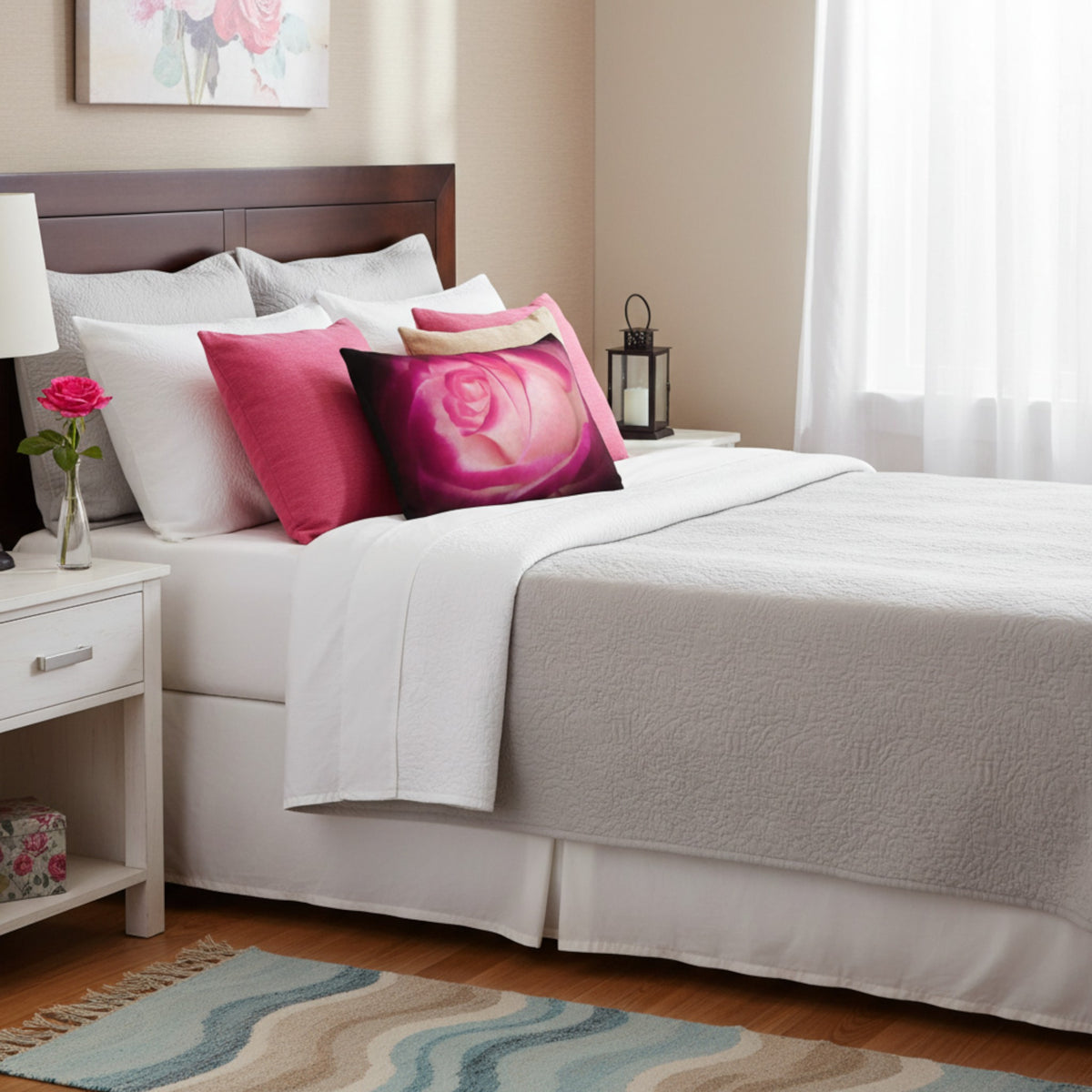 A neatly made bed in a bright bedroom setting, featuring white bedding, a textured light gray throw blanket, and several decorative pillows including a large pink rose print pillow and a solid magenta pillow.