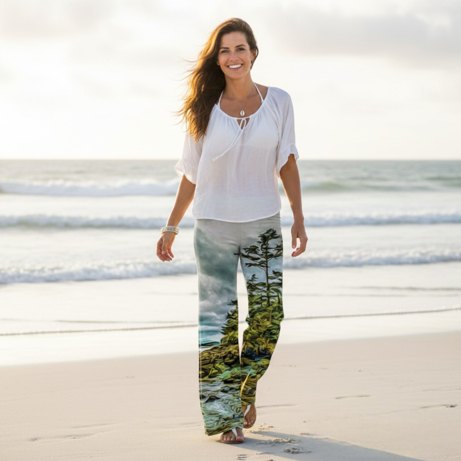 Woman walking on a beach wearing a white top and pants with a nature print.