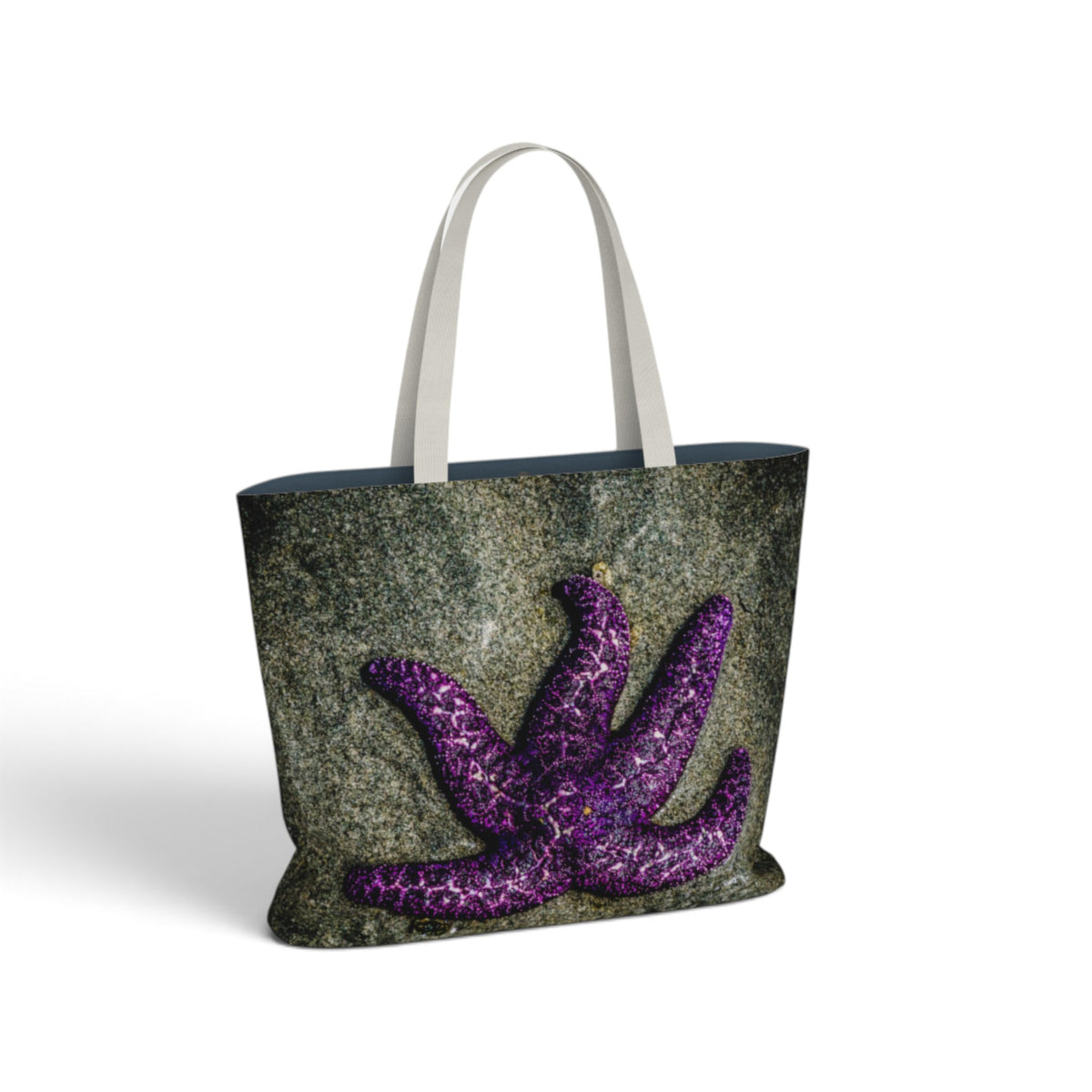 A tote bag featuring a textured dark gray and green stone-like print with a large purple starfish design on the front, finished with white shoulder straps.