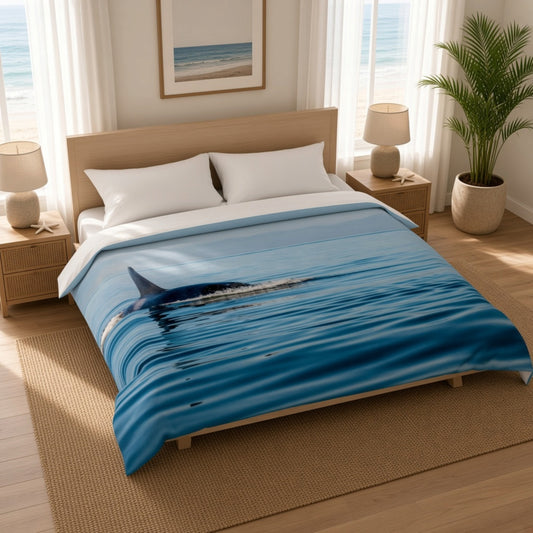 A bed dressed in a blue ocean-print comforter featuring a shark fin and rippling water, styled in a bright bedroom with wood furniture, lamps, a plant, and ocean-view windows.