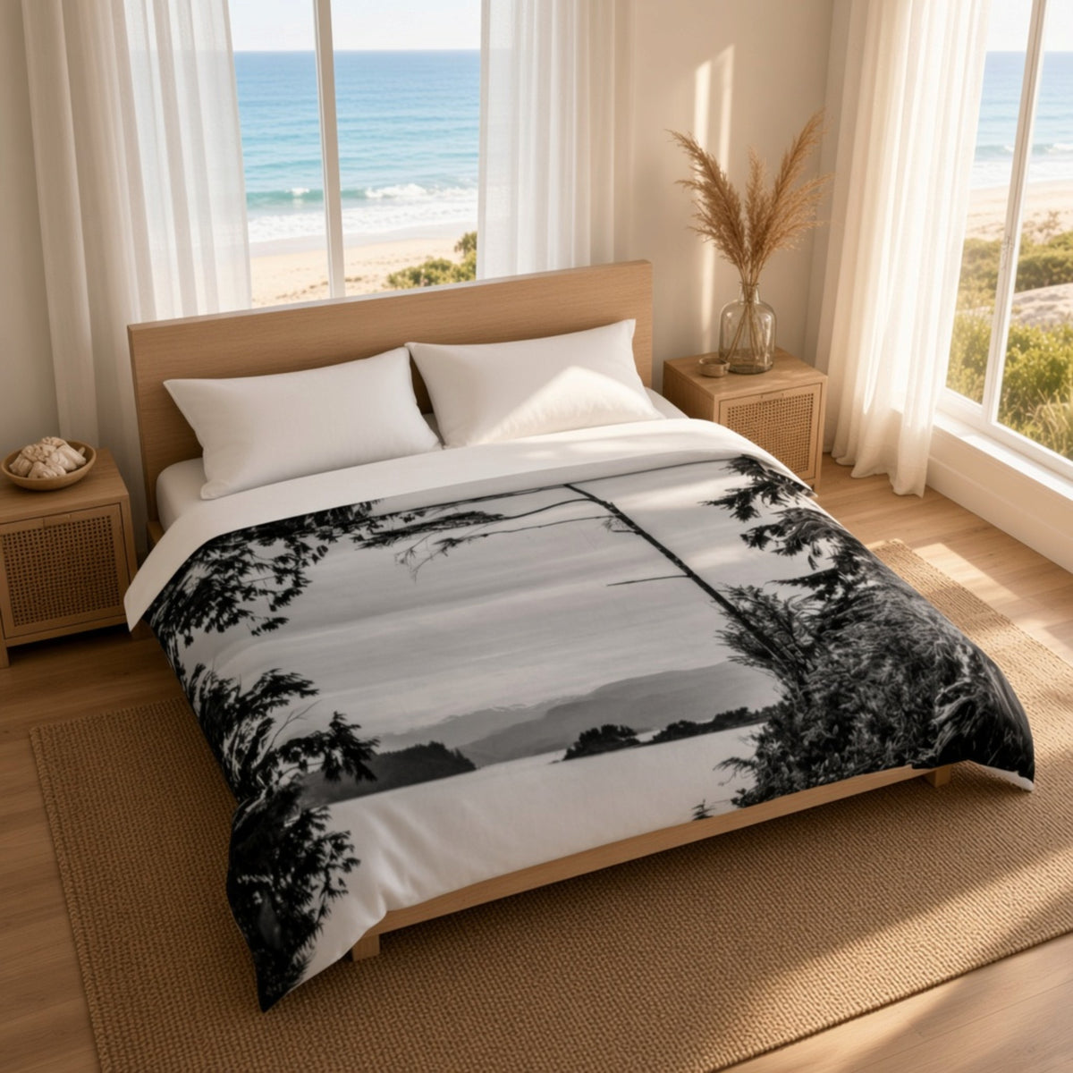 A bed dressed in a black-and-white scenic duvet or comforter with a monochrome landscape print, styled in a bright bedroom with wood furniture, sheer curtains, and large windows overlooking the ocean.