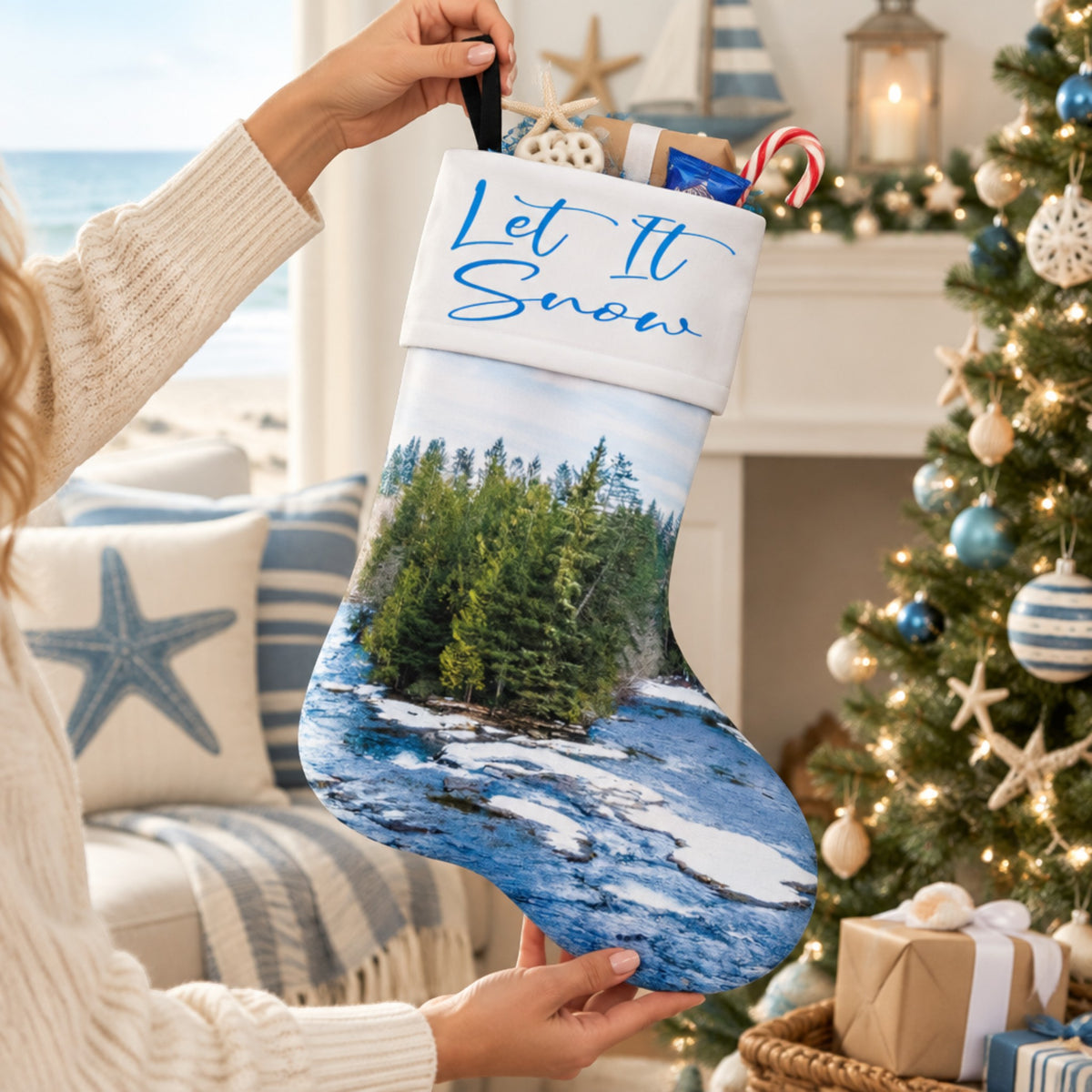 A person holds up a Christmas stocking featuring a printed winter landscape with blue snow, green trees, and white rocks. The stocking has a white cuff with blue script text reading 'Let It Snow.